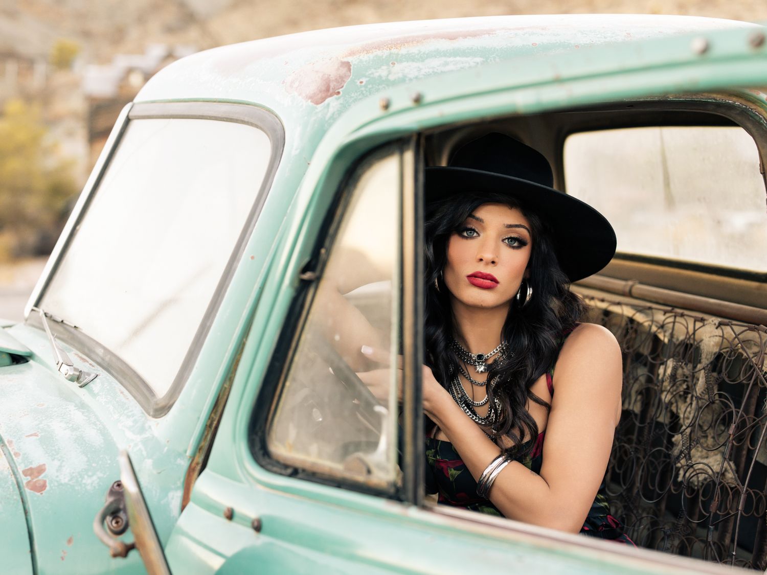 Woman in black outfit and wide-brim hat poses inside a vintage turquoise pickup truck in the desert.