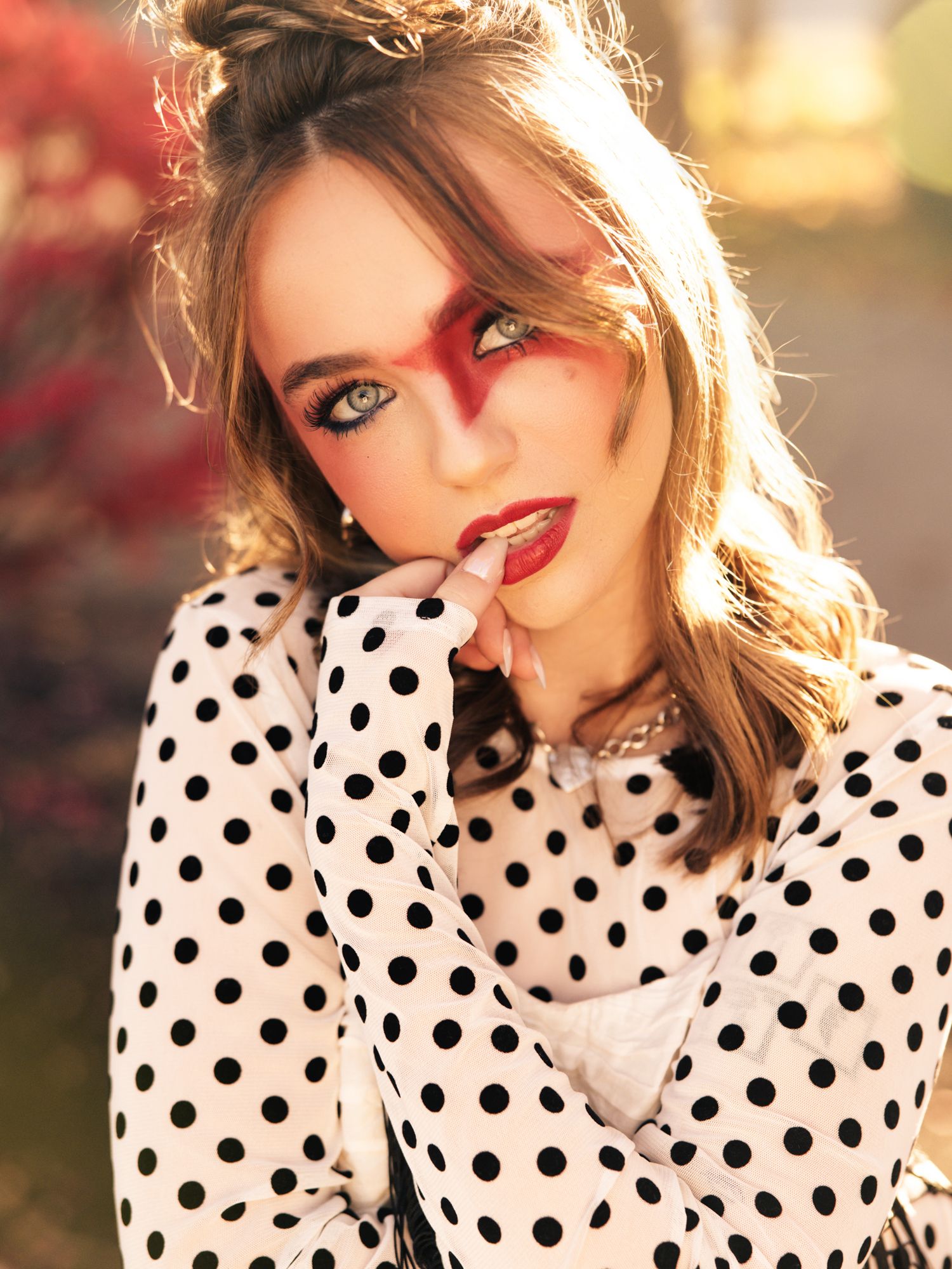 Woman in black and white polka dot blouse with dramatic red makeup poses outdoors in warm sunlight.