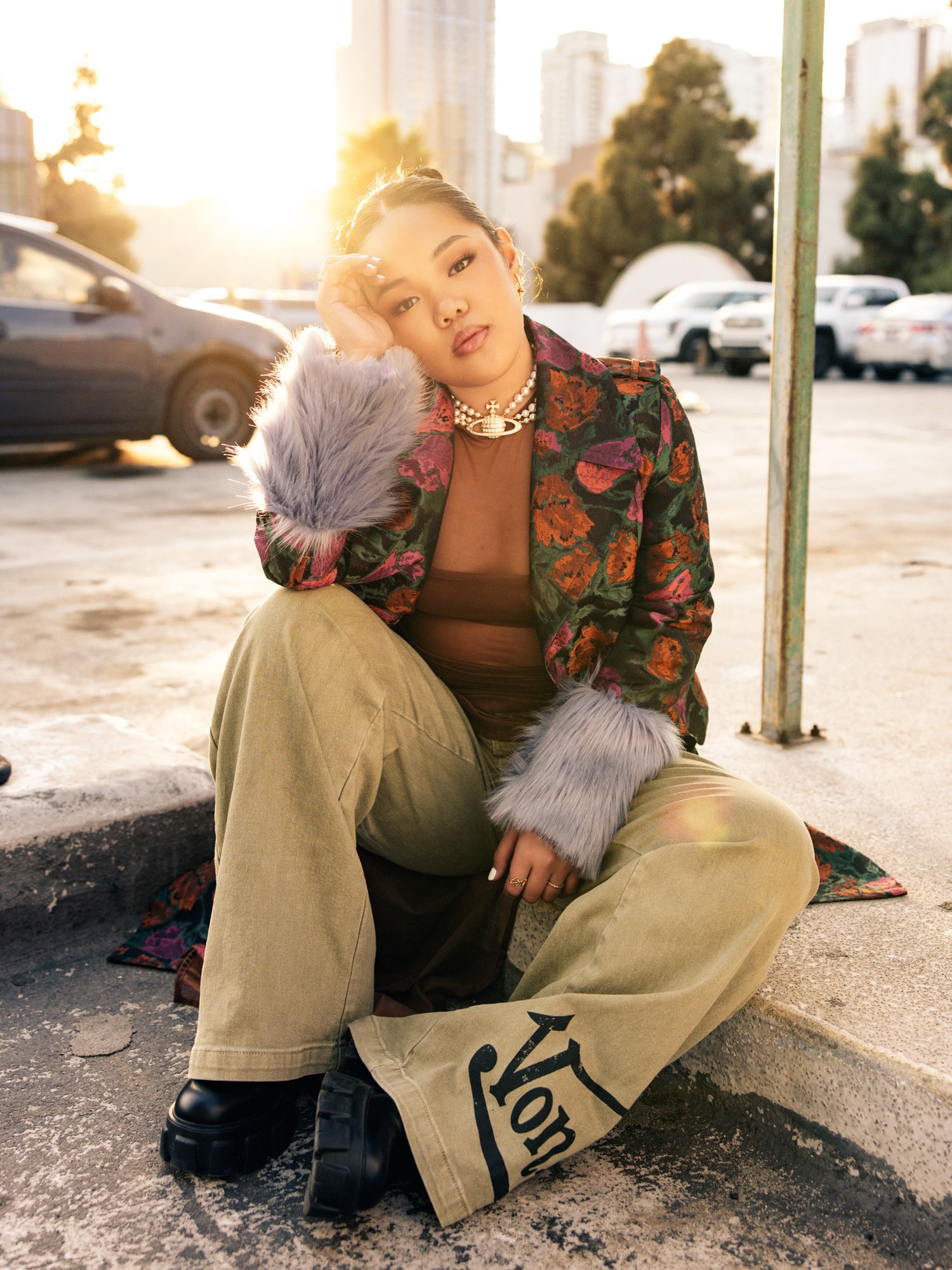 Woman in floral jacket with fur trim and wide-leg pants sitting outdoors at golden hour.
