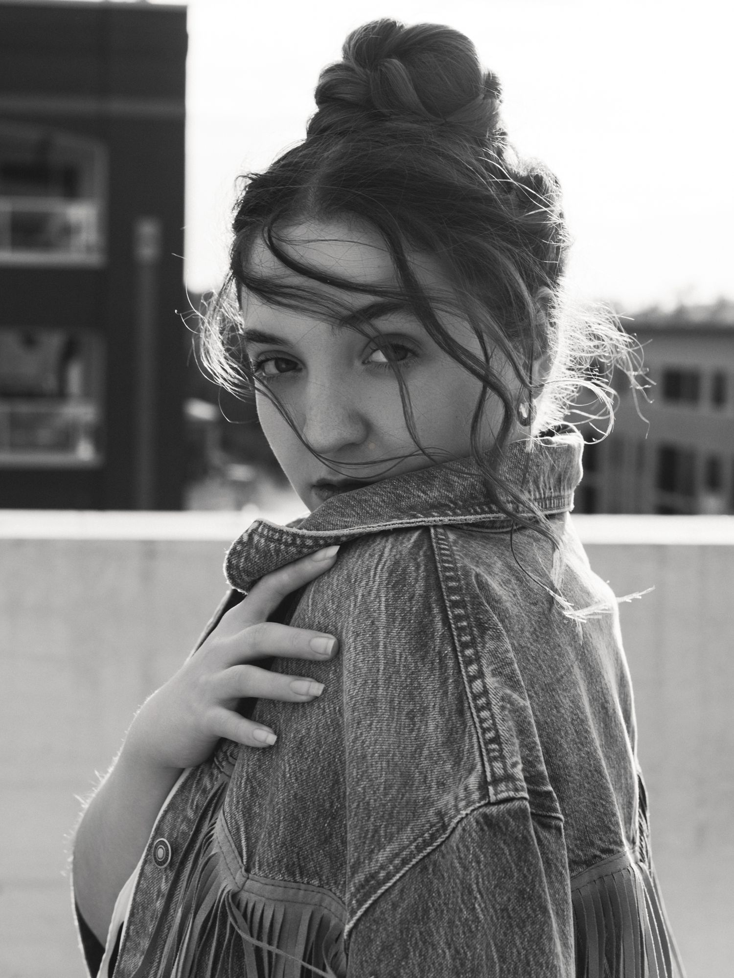 Black and white portrait of a young woman with a bun updo wearing a denim jacket on a rooftop.