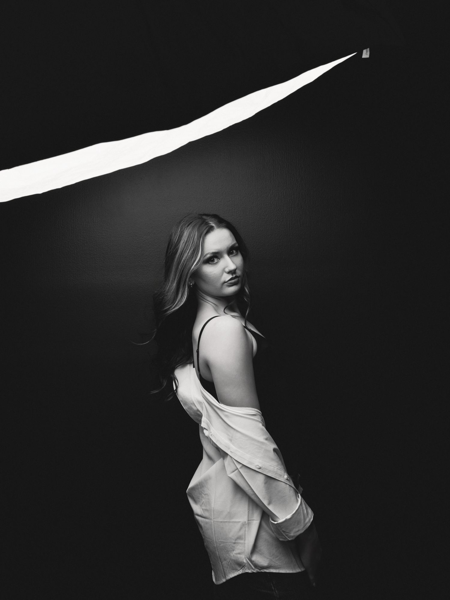 Black and white photo of a woman in a white top glancing back over her shoulder in dramatic lighting.