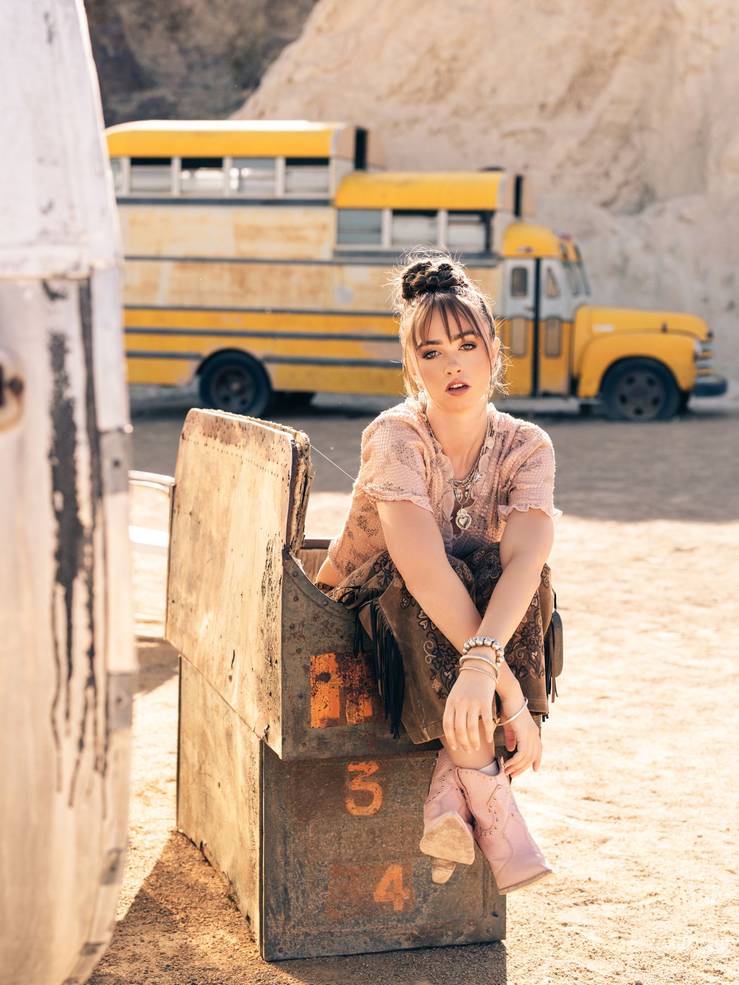 Young woman in floral outfit sits on wooden crates in a dusty lot with yellow school buses behind her.