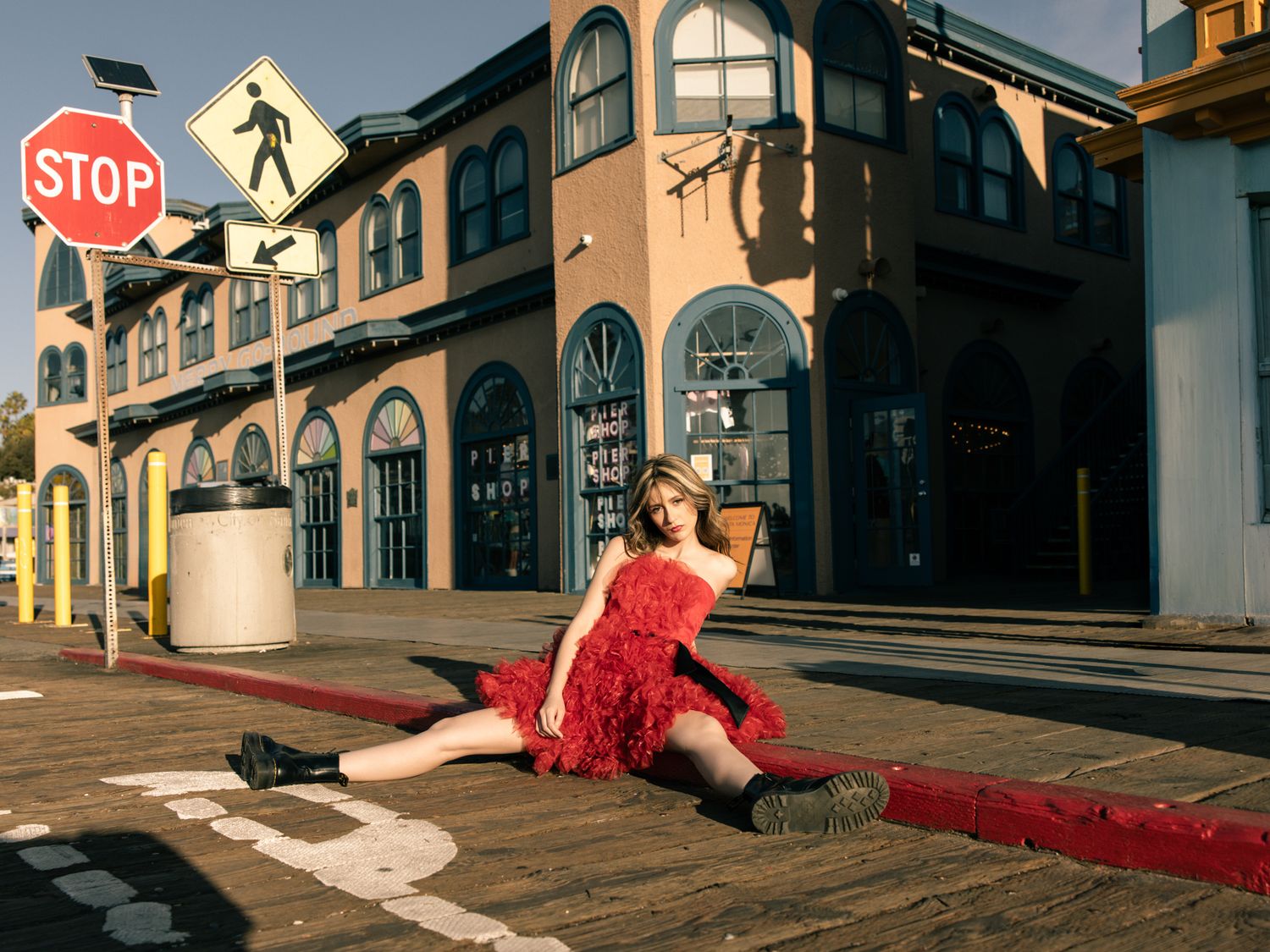 Woman in red ruffled dress doing splits on wooden boardwalk near stop sign at sunset.