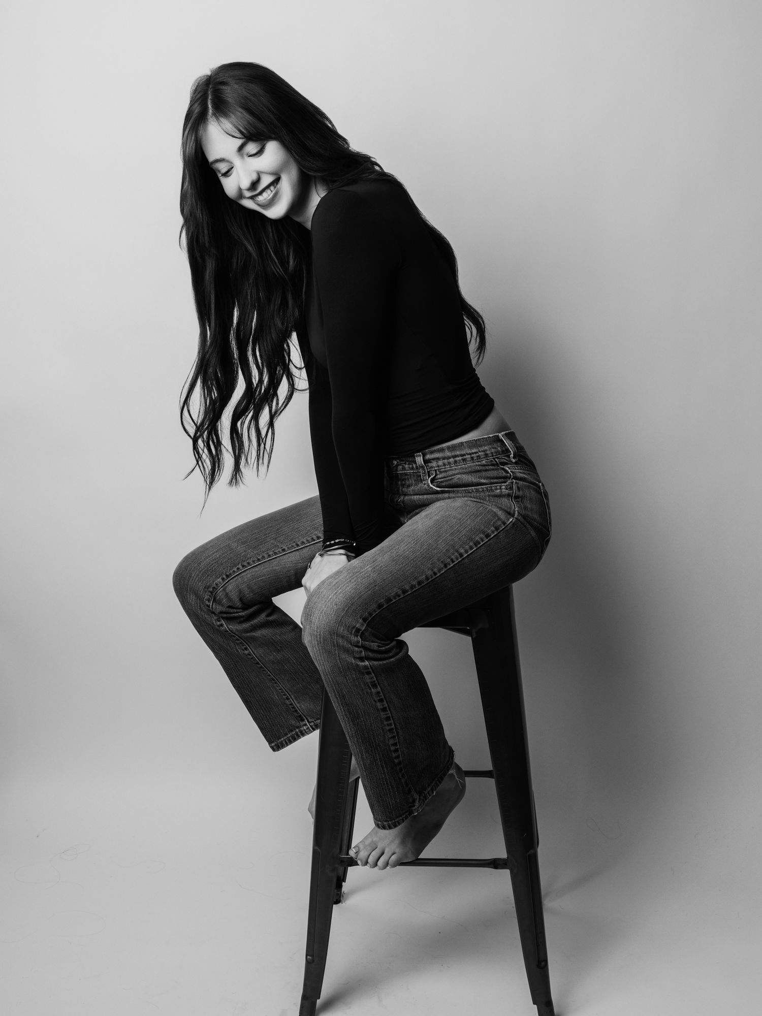 Black and white photo of a young woman sitting on a stool, wearing jeans and black top, smiling and looking down.