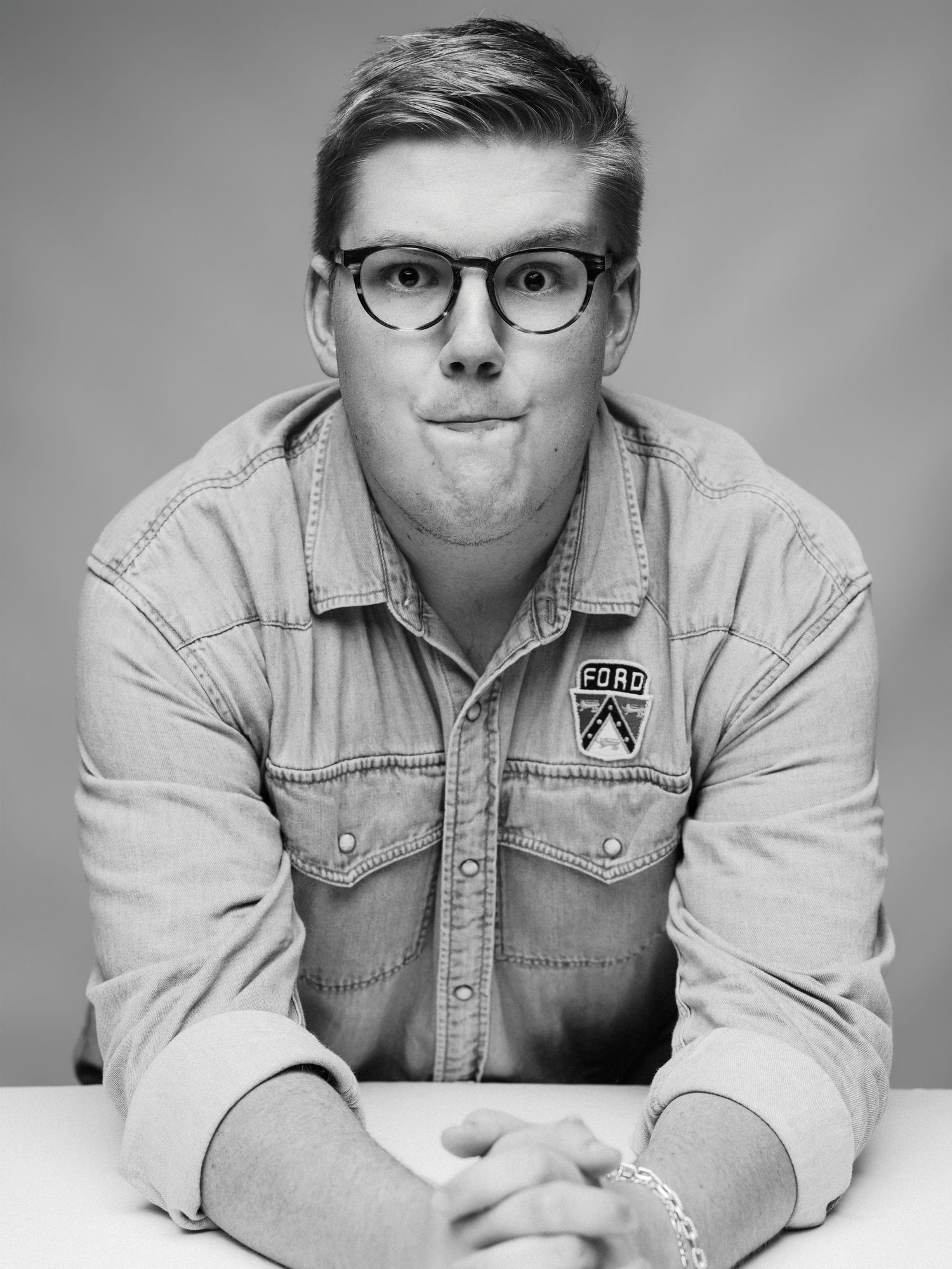 Black and white photo of a young man in glasses and a Ford patch denim shirt making a funny face.