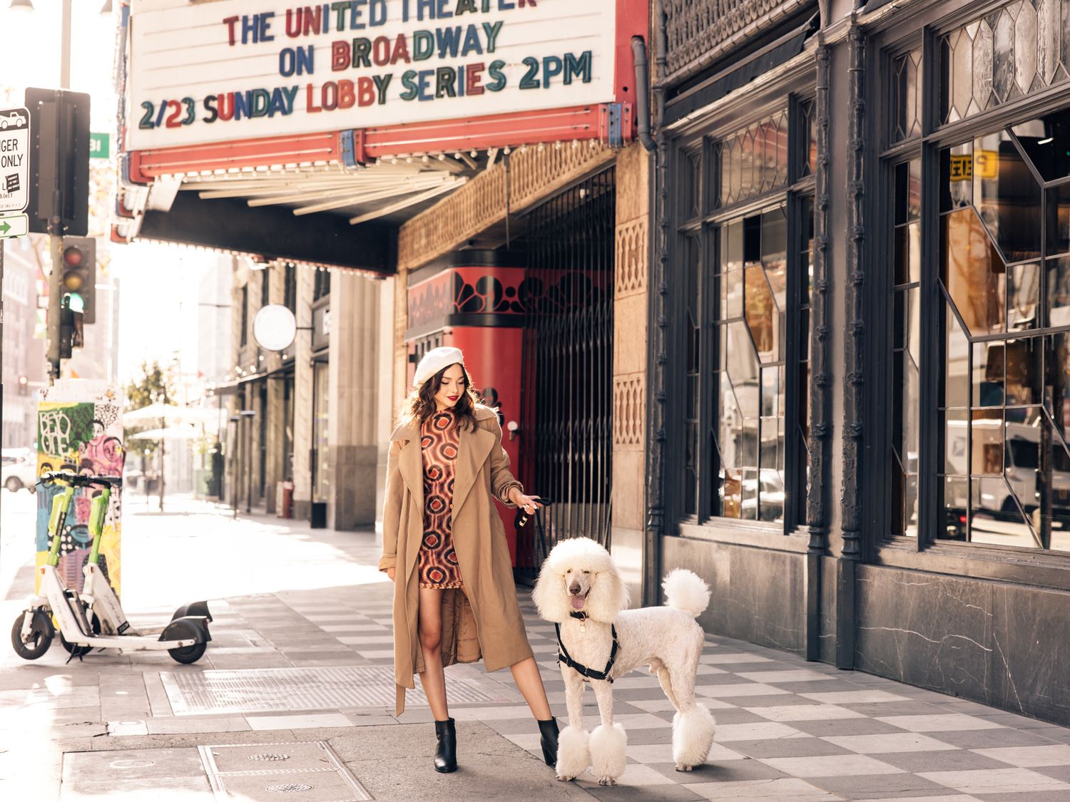 Fashion model in camel coat walking white poodle outside United Theater on Broadway street.