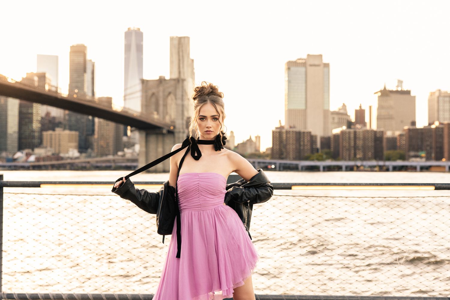 Woman in pink mini dress and leather jacket posing by river with NYC skyline at sunset.