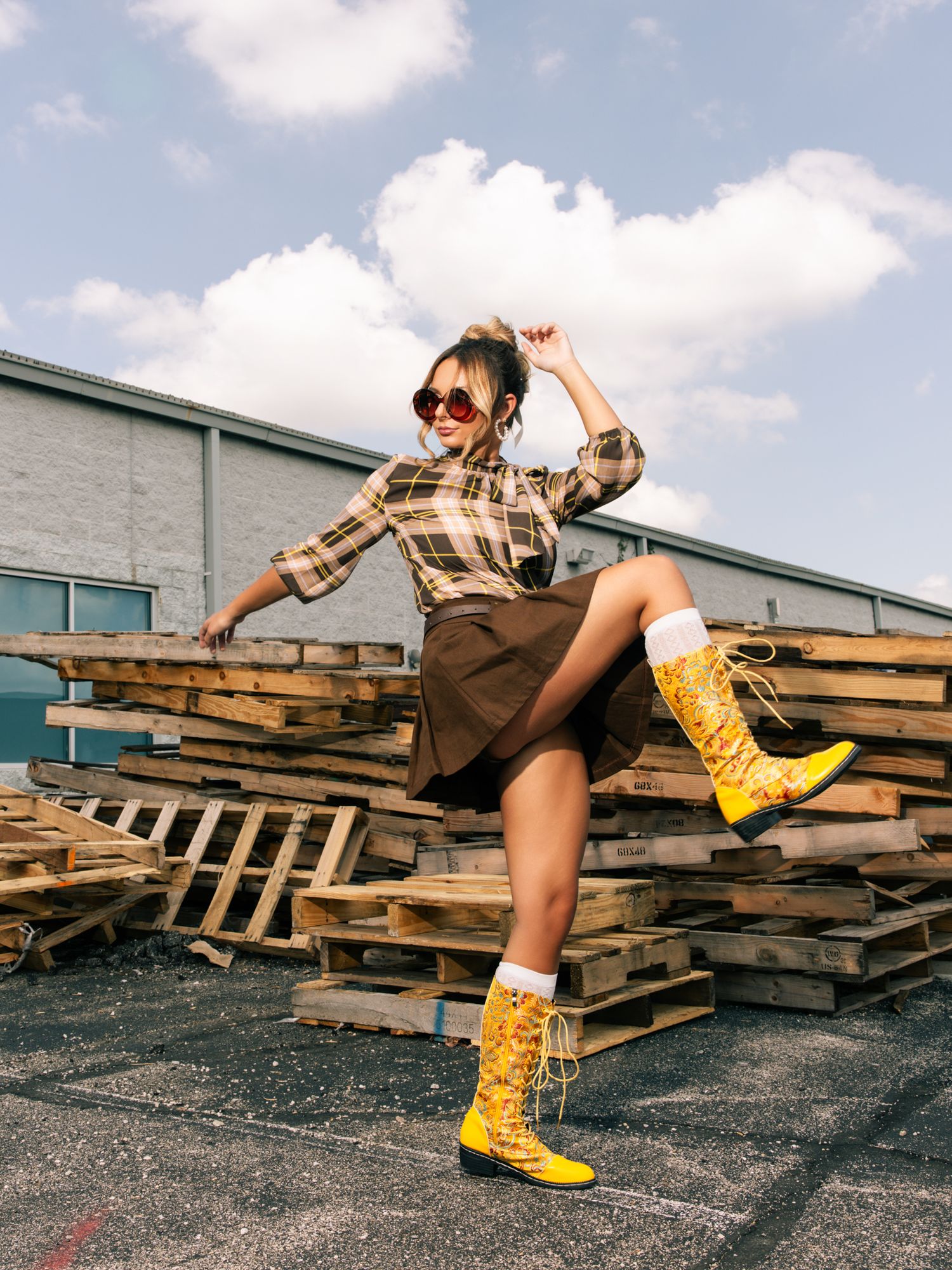 Woman in plaid shirt and brown skirt posing with yellow boots on wooden pallets outdoors.