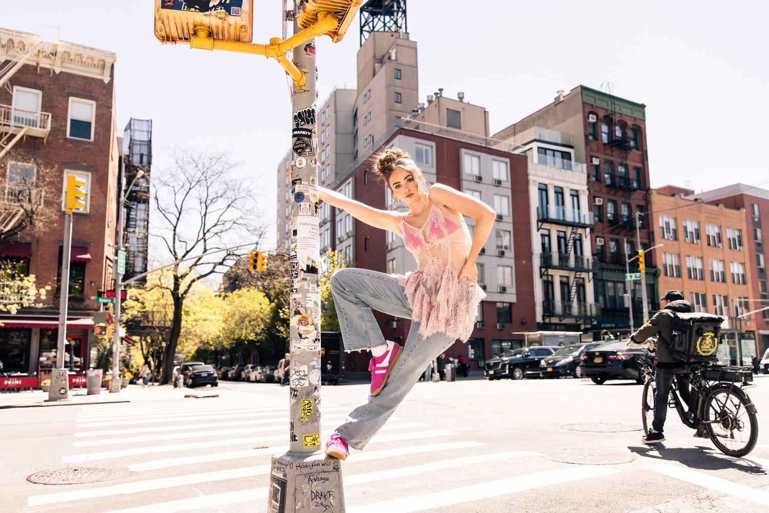 Woman in pink top and gray pants posing on street pole at NYC intersection.