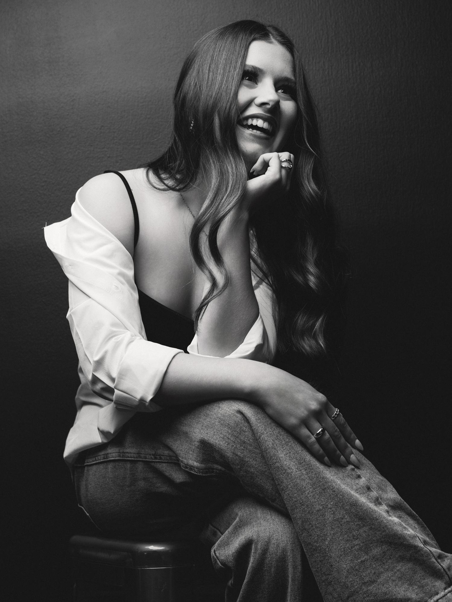 Black and white photo of smiling woman with long hair sitting on a stool in a white shirt and jeans.