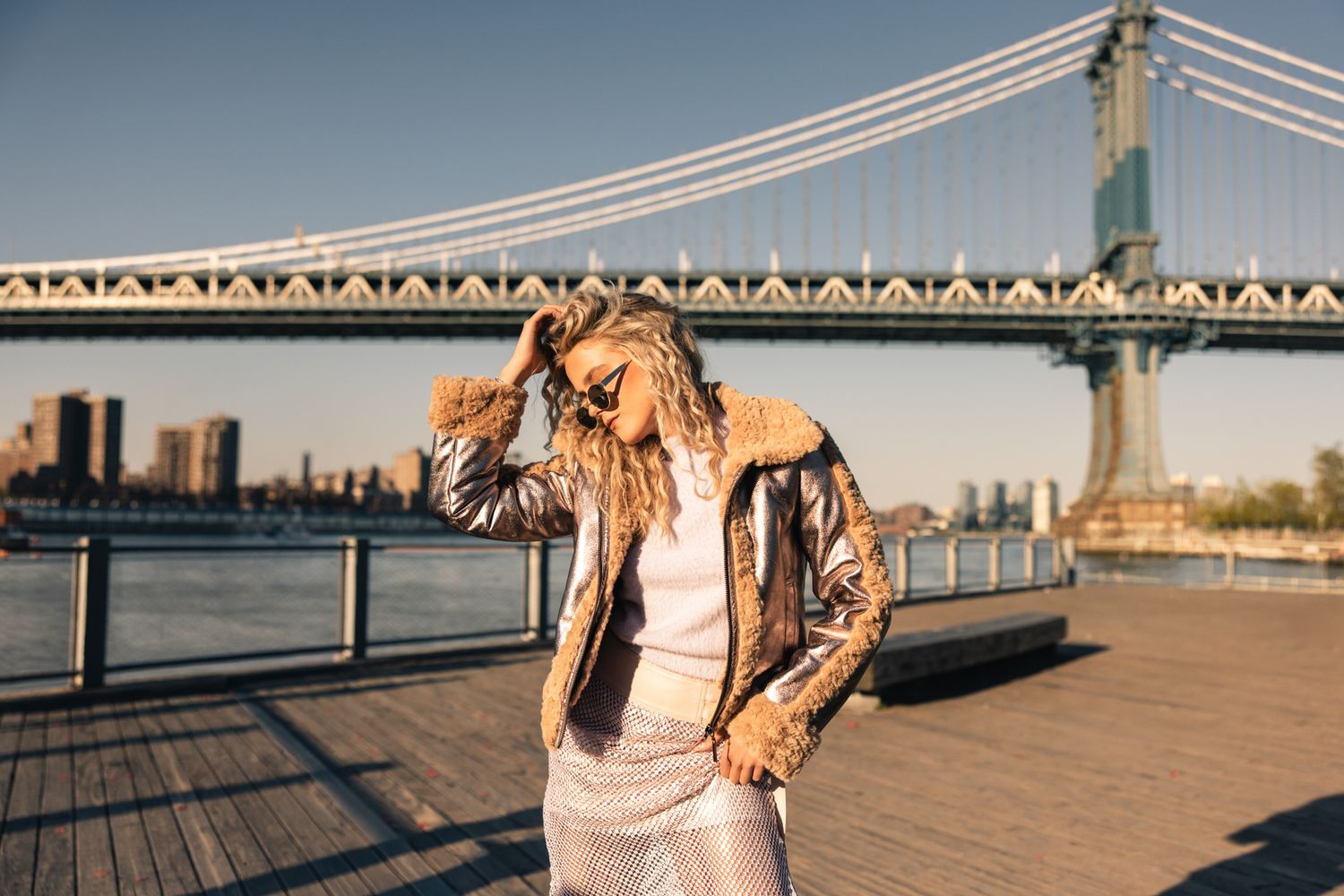 Woman in metallic jacket posing on waterfront boardwalk with Manhattan Bridge behind her.
