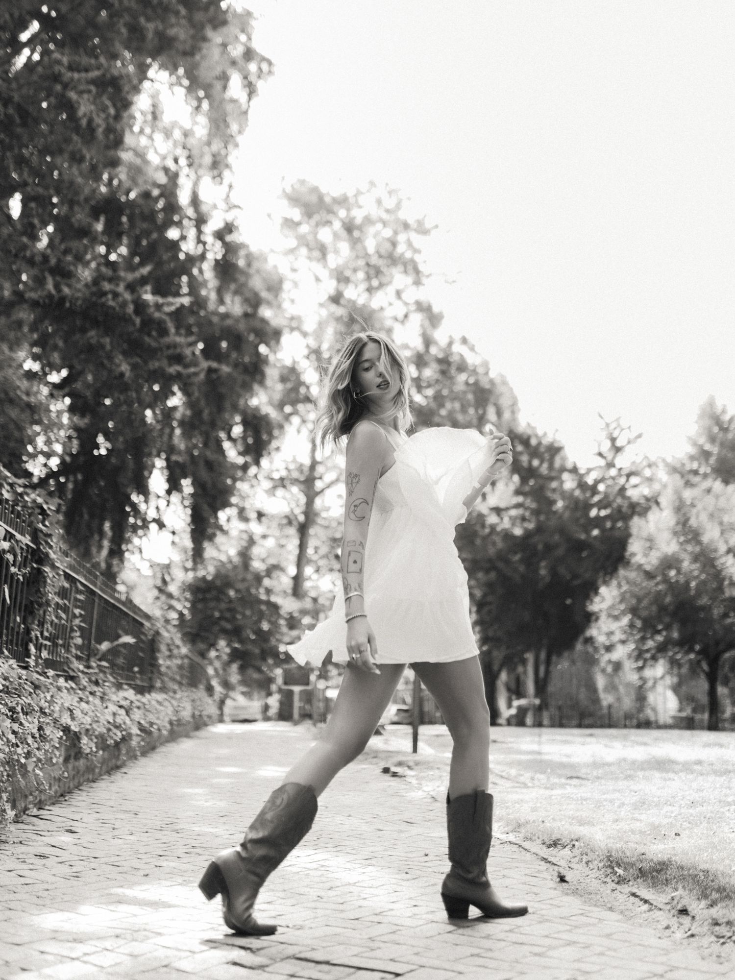 Black and white photo of a woman in a white mini dress and cowboy boots posing on a tree-lined brick path.
