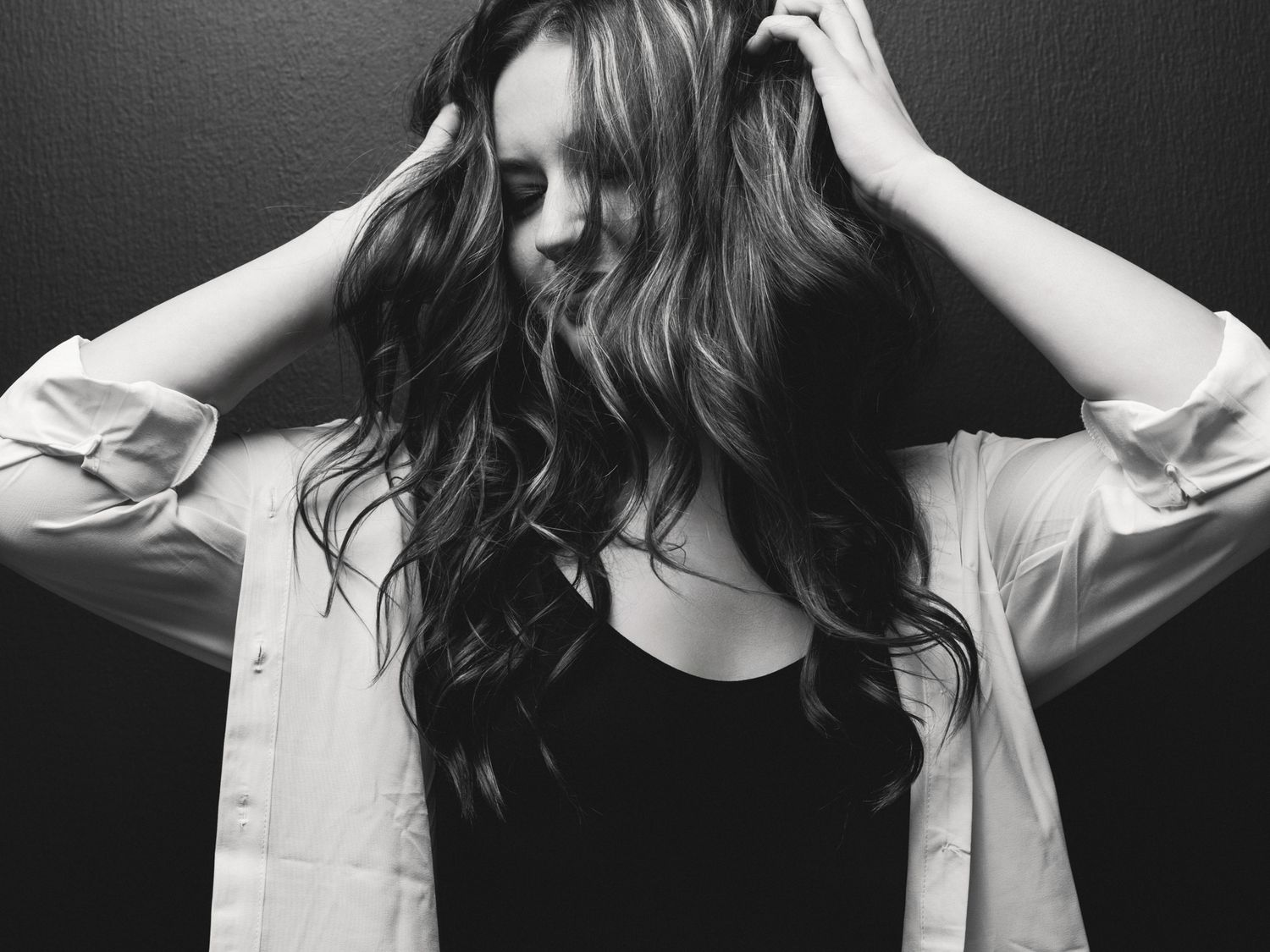 Black and white portrait of a woman with wavy hair running her hands through it against a dark background.