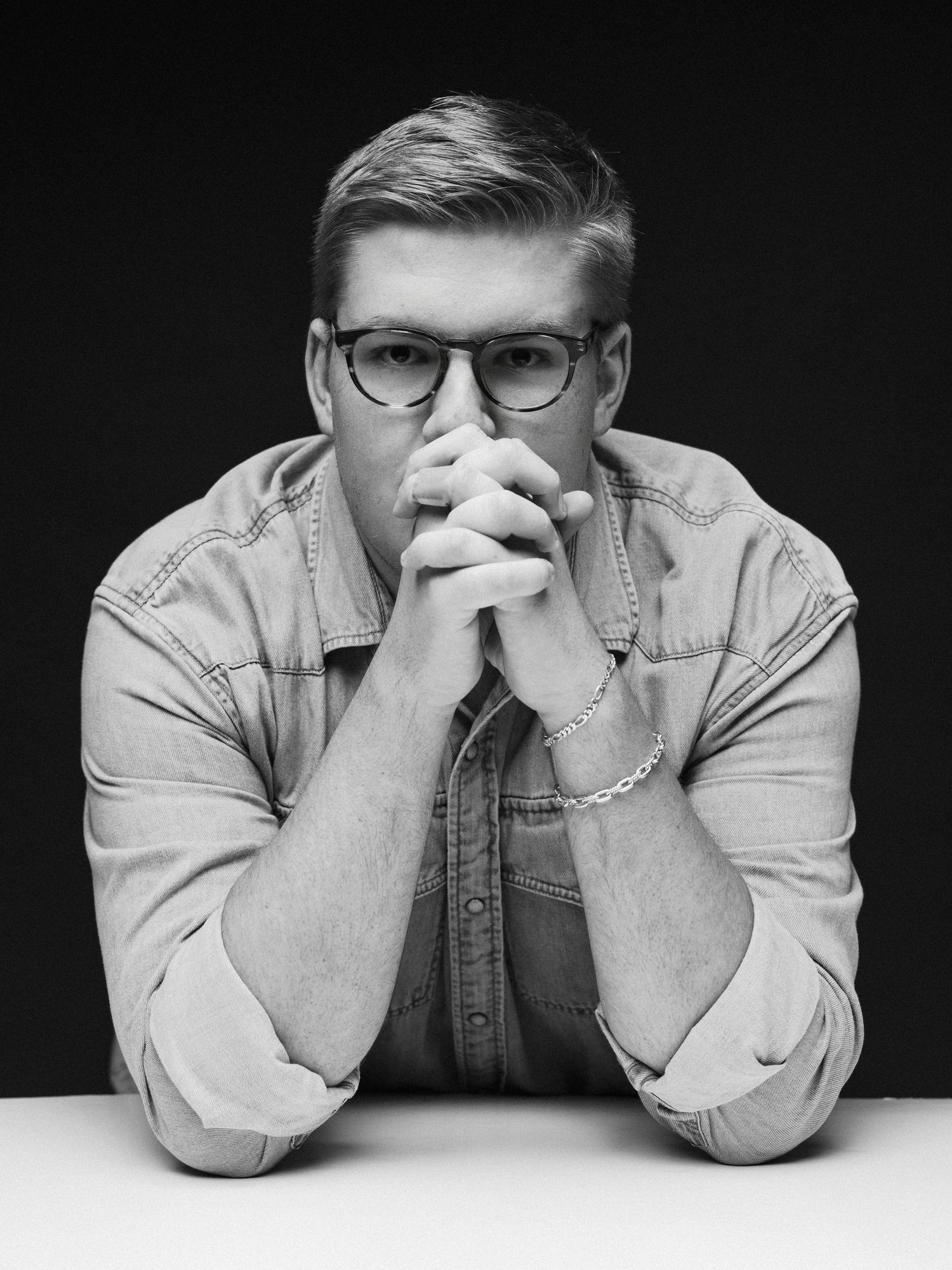 Black and white portrait of a young man with glasses and a denim shirt with hands clasped at a table.