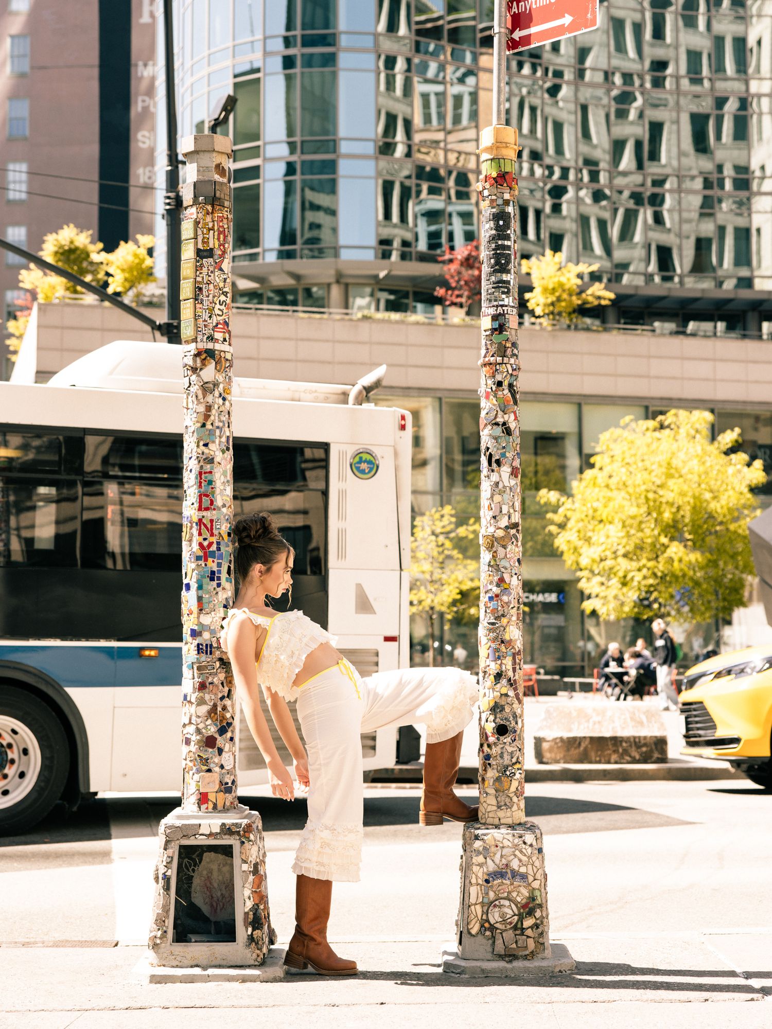 Woman in white outfit with brown boots posing between decorated poles at urban city bus stop.