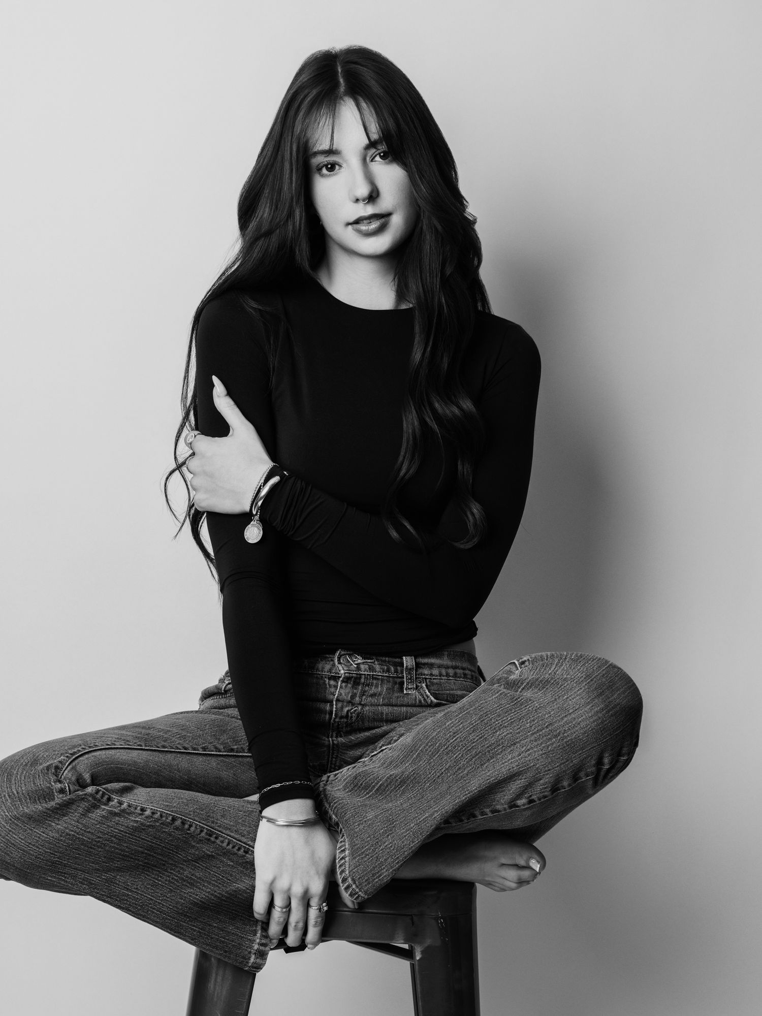 Black and white portrait of a young woman in black top and jeans sitting on a stool with crossed legs.