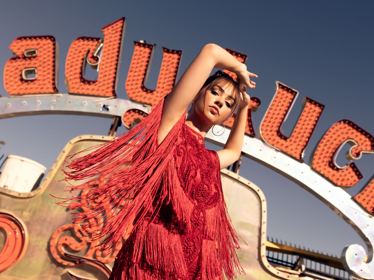 Woman in red fringe dress poses with arms raised beneath a colorful Lady Luck neon sign outdoors.