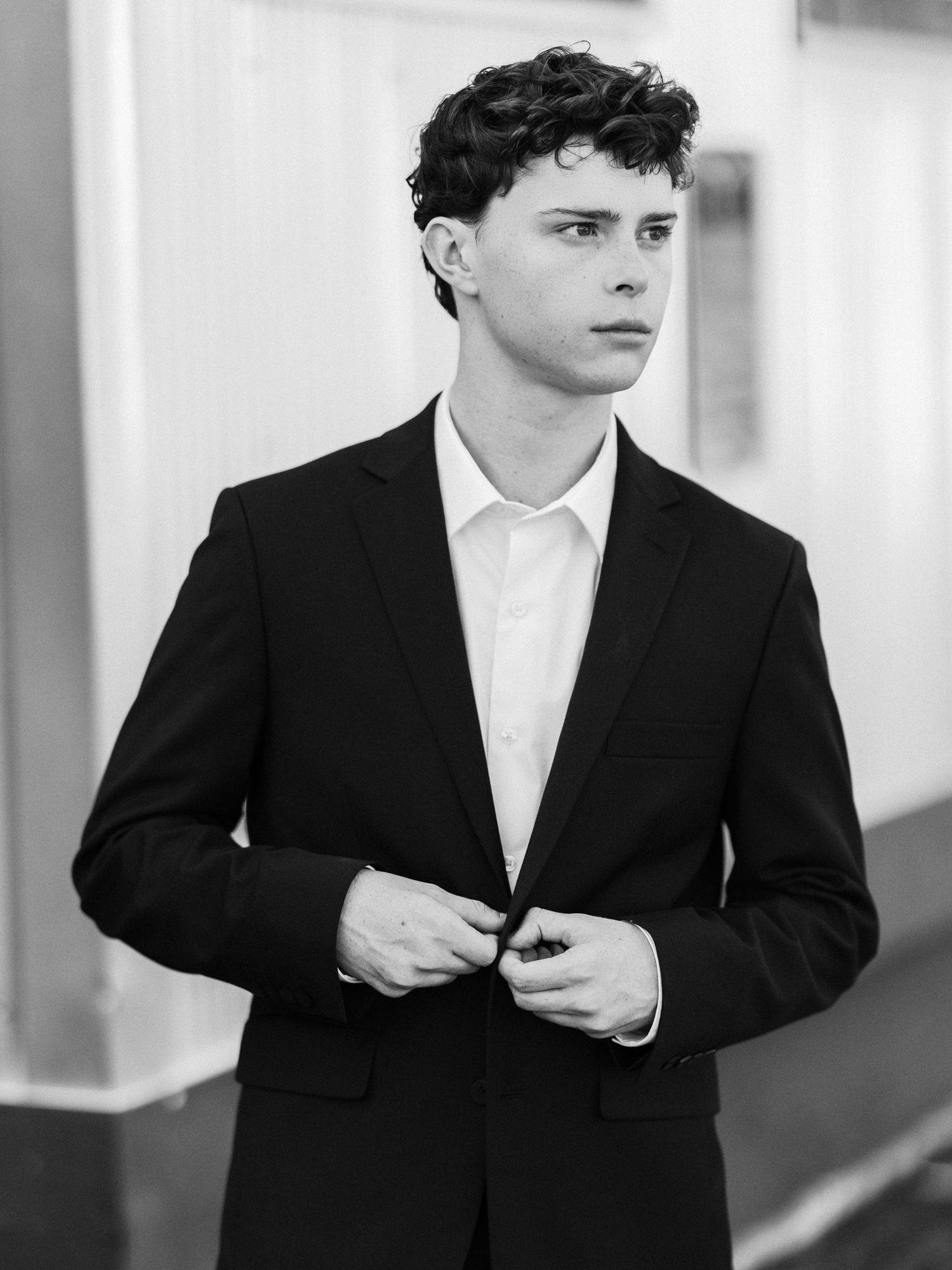 Black and white portrait of young man in black suit adjusting jacket buttons outdoors.