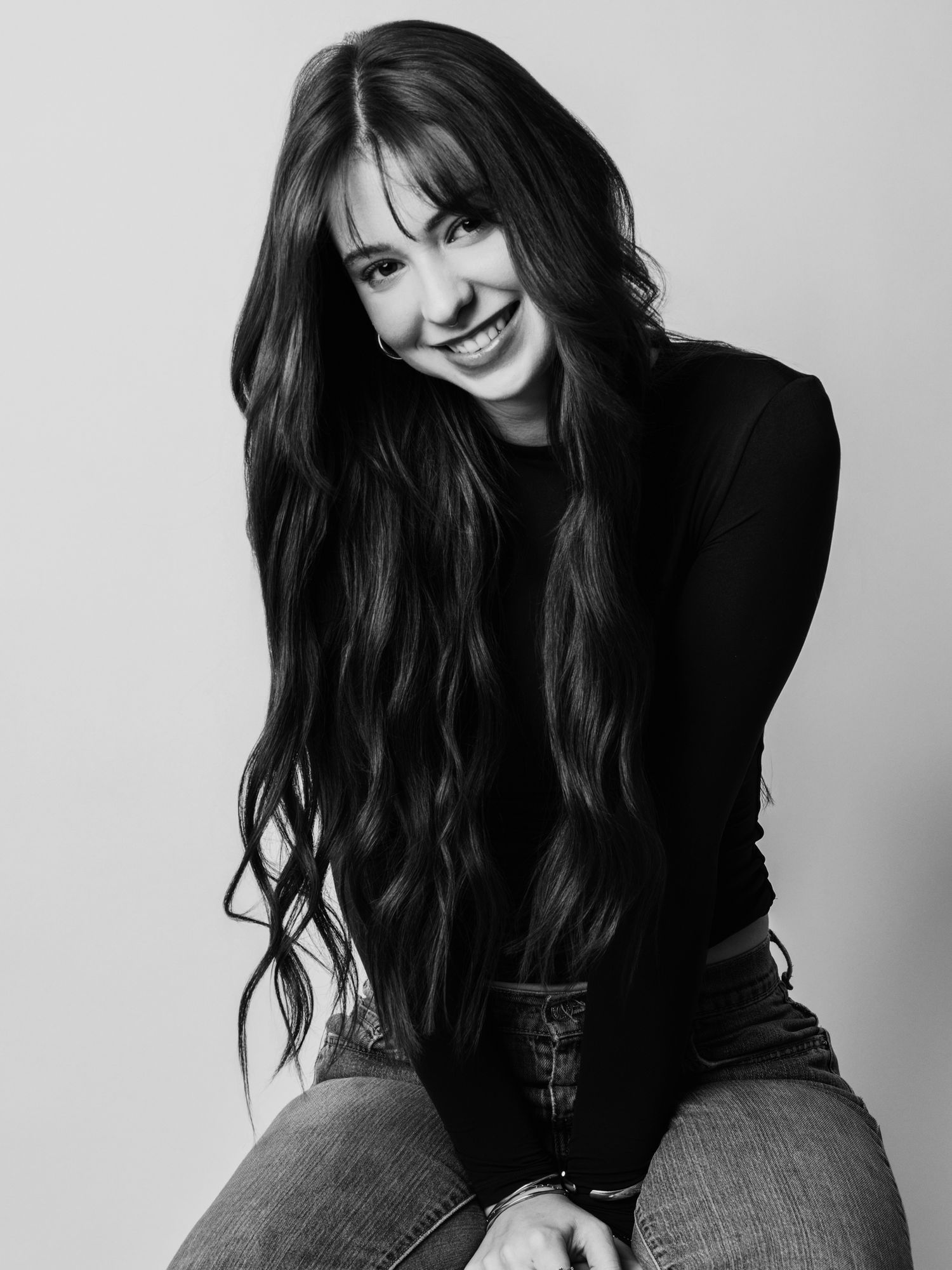 Black and white portrait of a smiling young woman with long wavy hair wearing a black top and jeans.