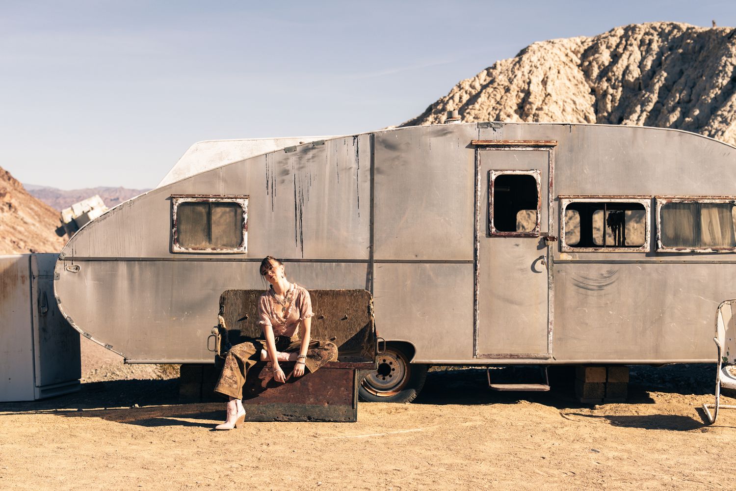 Person sitting on steps of a weathered vintage silver trailer in an arid desert landscape.