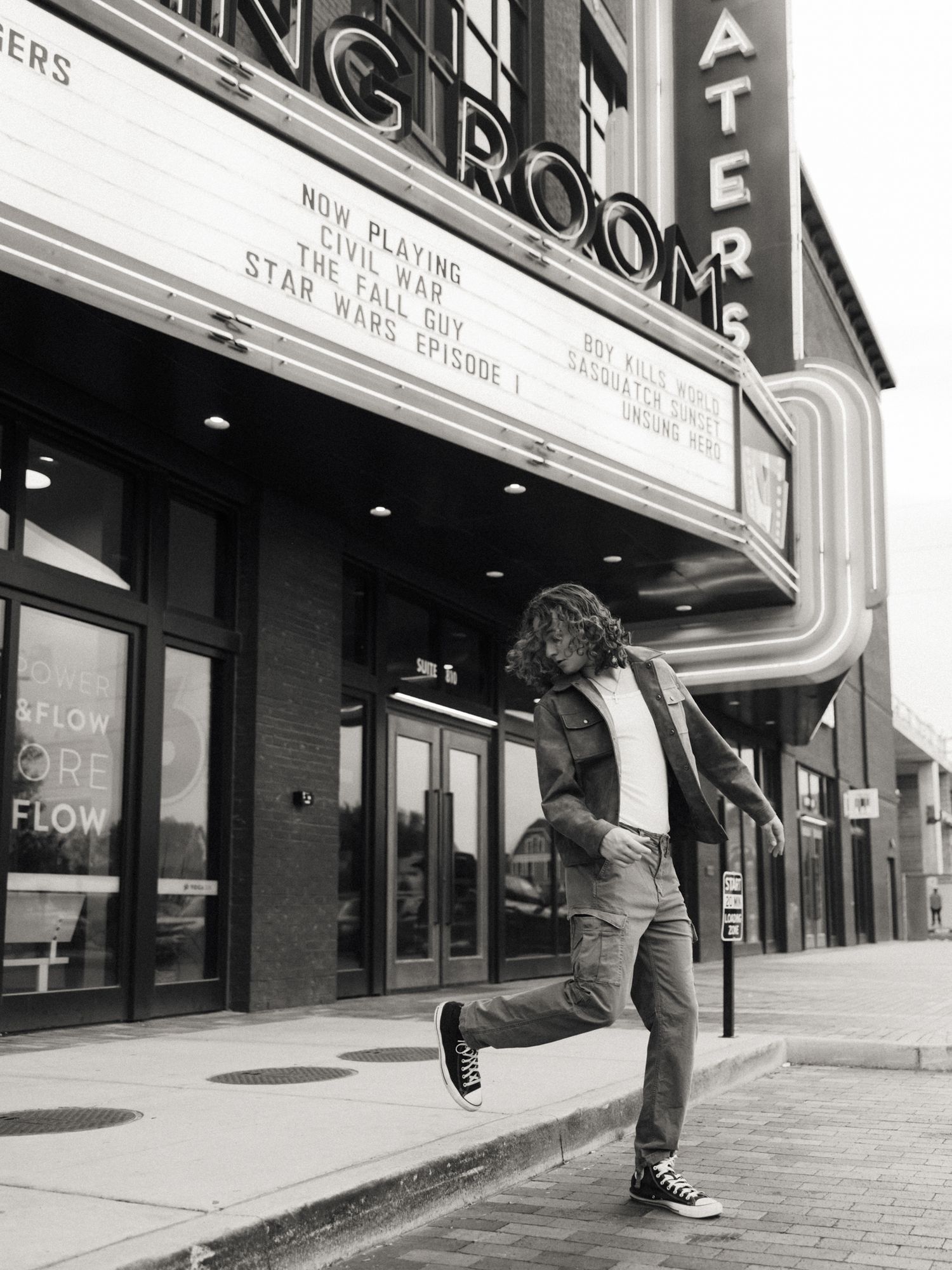 Black and white photo of a person dancing outside a classic movie theater marquee on a city sidewalk.