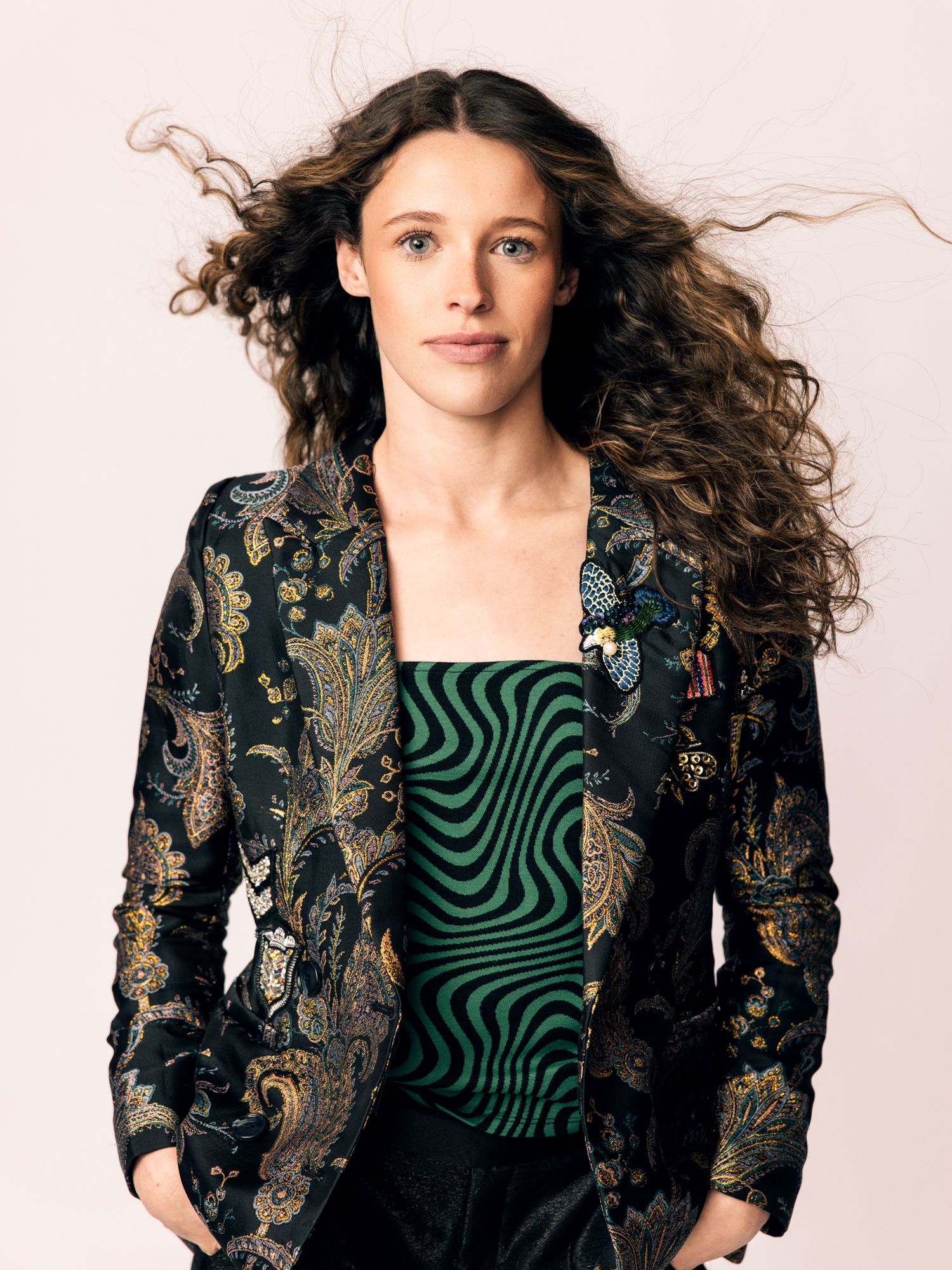 Woman with long curly hair in floral blazer and green striped top posing against light background.