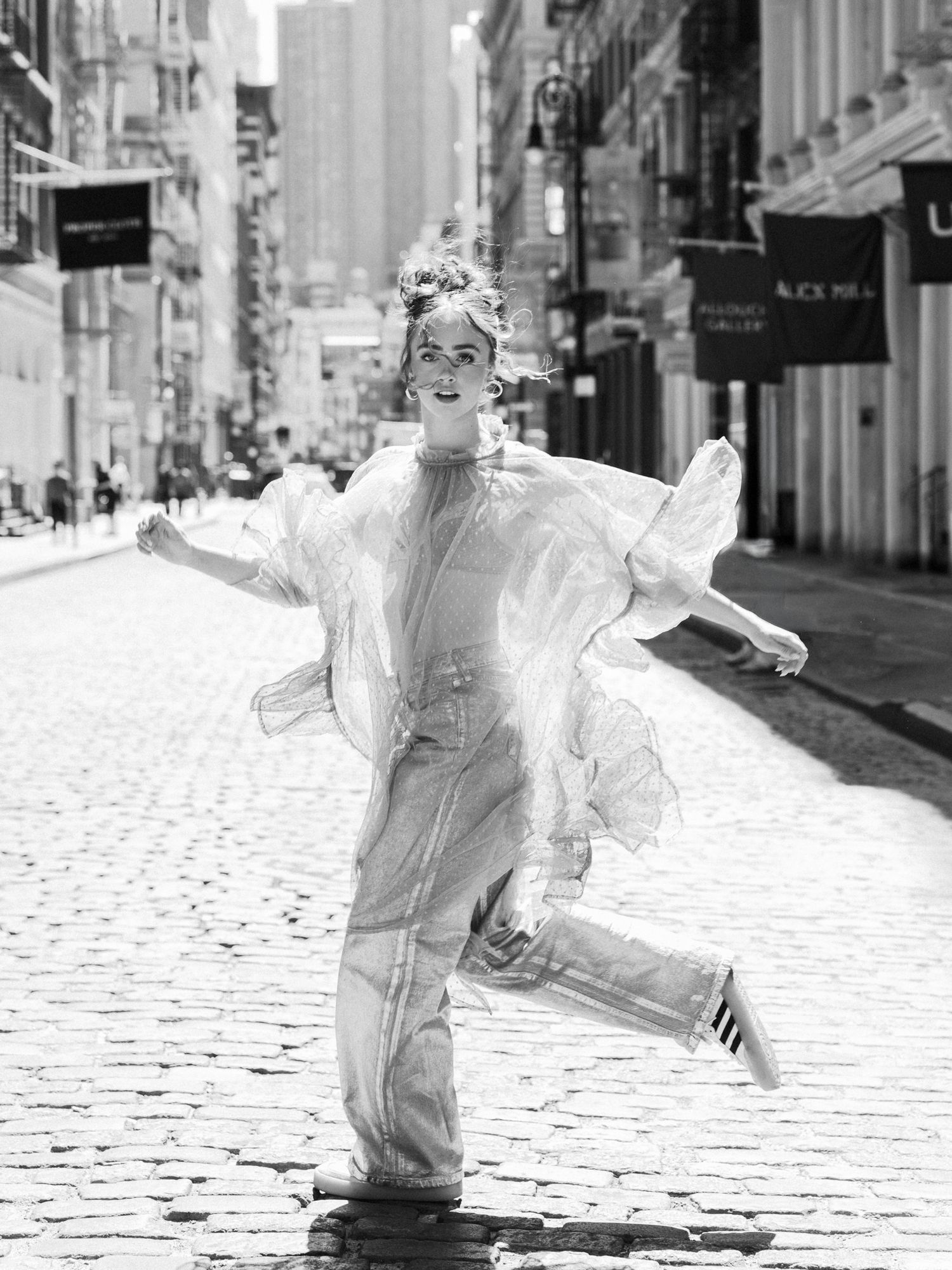 Black and white photo of a woman dancing in sheer outfit on cobblestone NYC street.