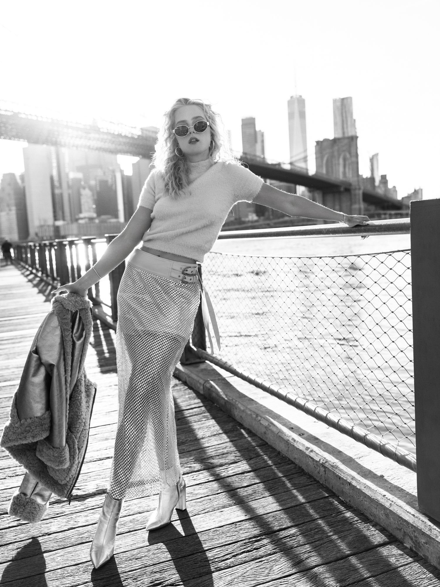 Black and white photo of blonde woman in checkered pants posing on Brooklyn Bridge boardwalk.