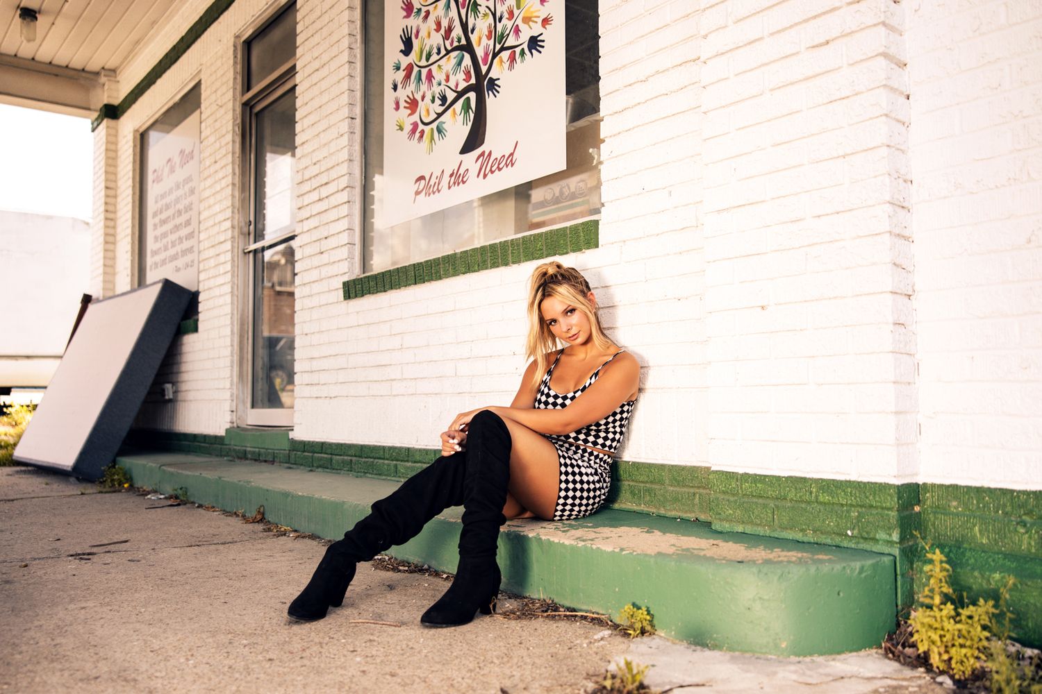 Blonde woman in houndstooth mini dress and black thigh-high boots sitting on steps outside a white brick building.