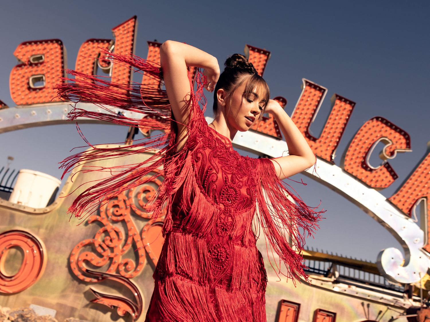Woman in red fringe dress poses dramatically beneath a vintage Lady Luck neon sign outdoors.