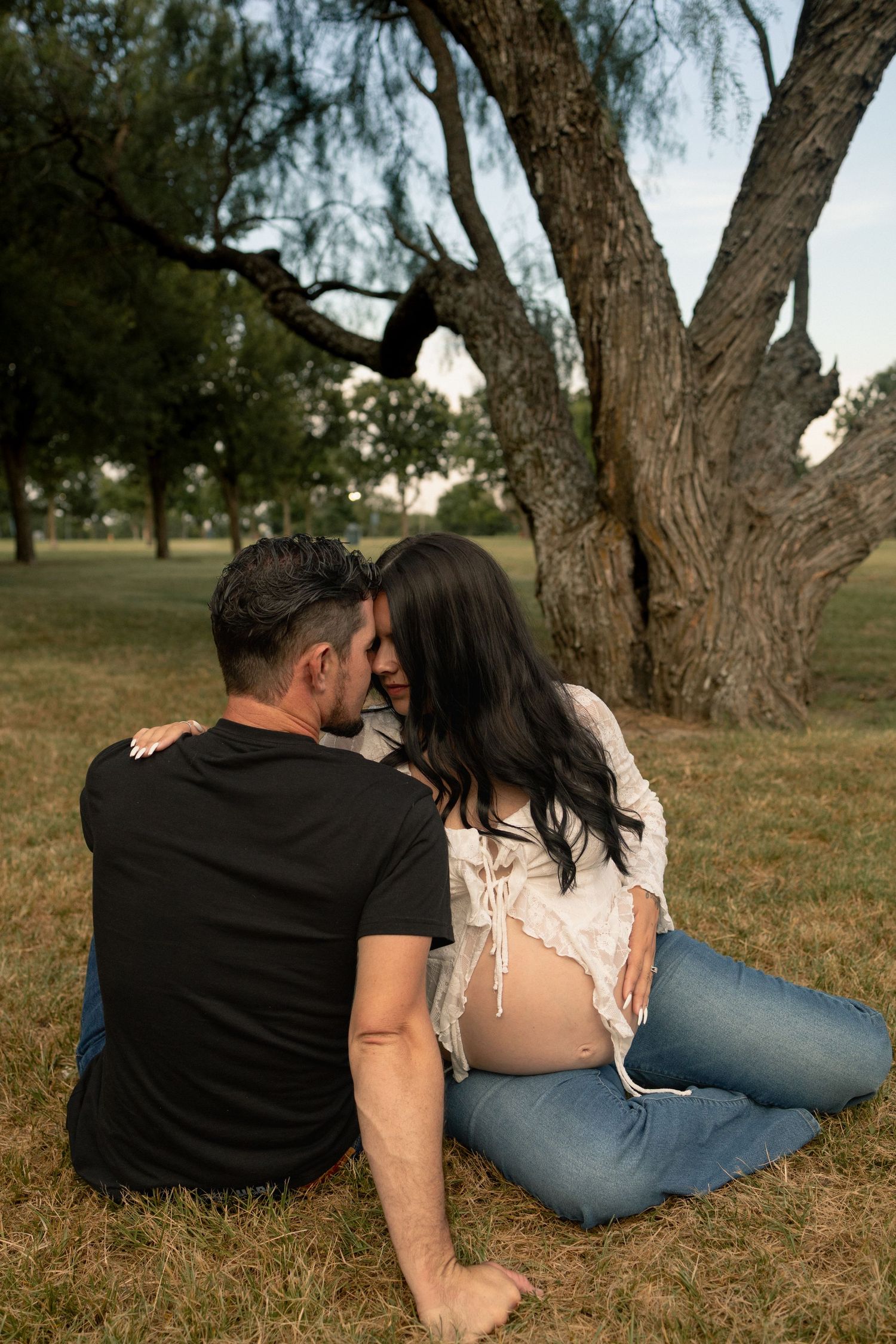 Couple sitting together on grass beneath a large tree, woman showing a pregnant belly in a park.