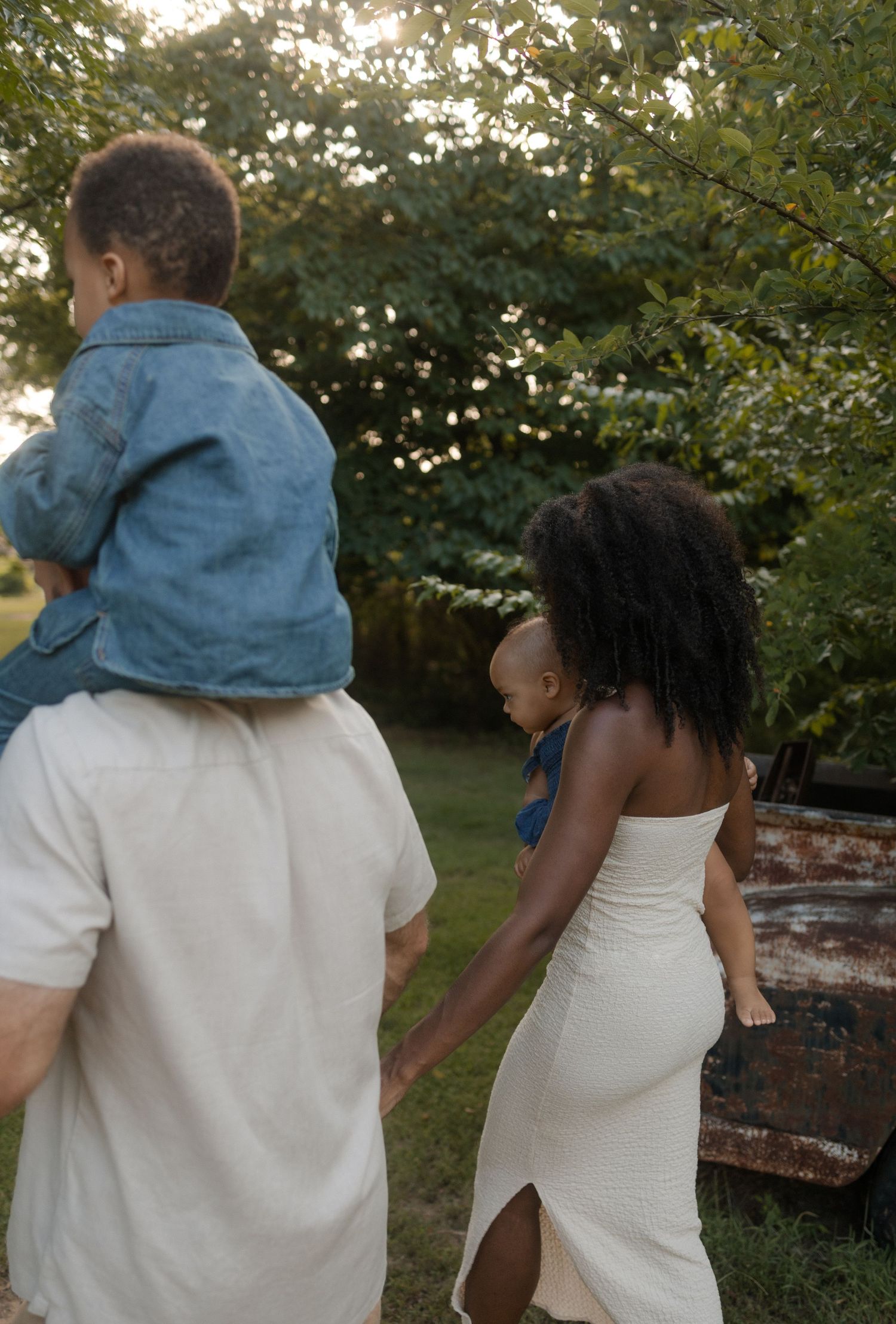 A family walks through a lush garden at sunset, father carrying toddler on shoulders while mother holds baby.