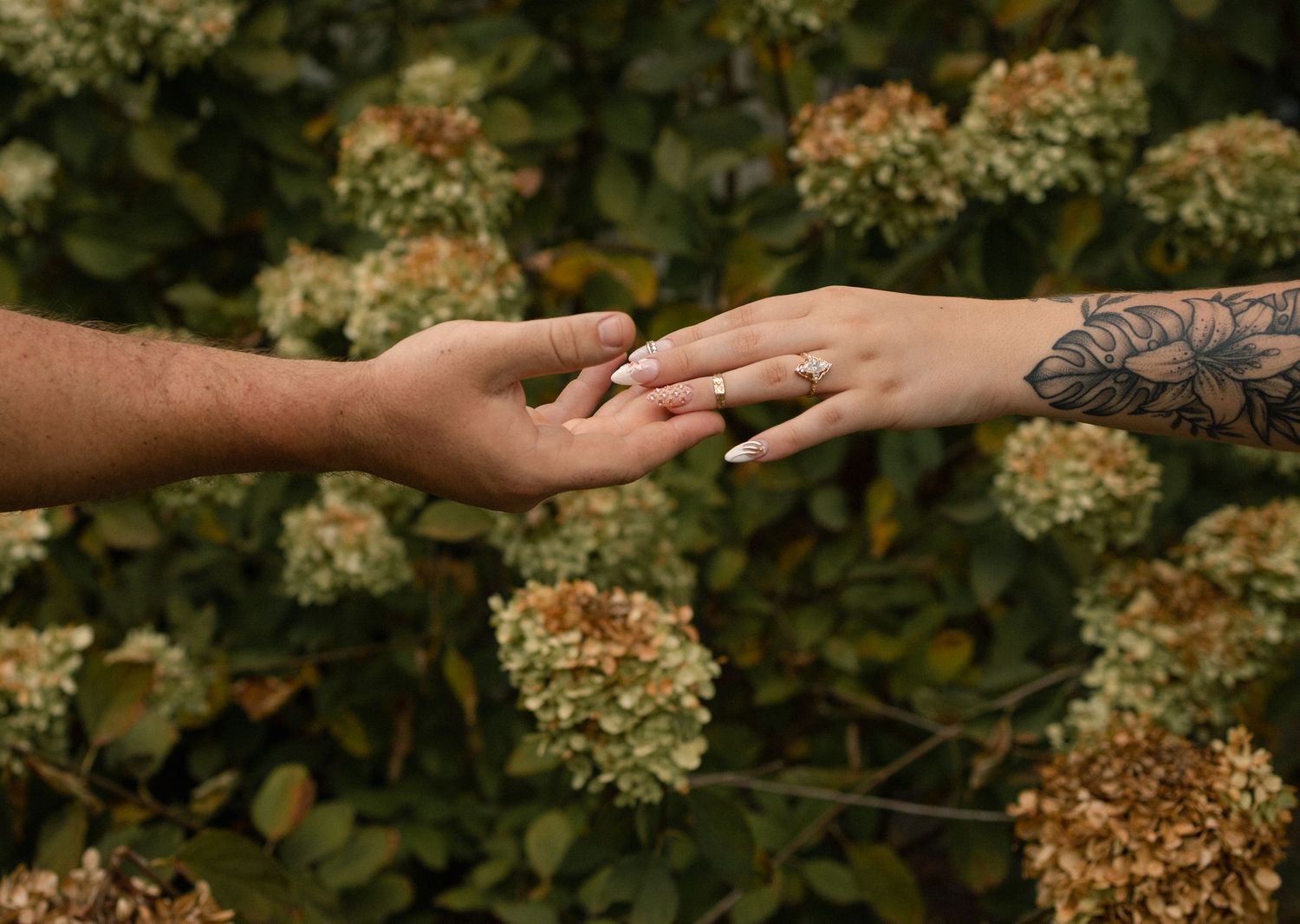 Couple's hands touching gently, woman wearing an engagement ring with a floral tattoo on her arm.