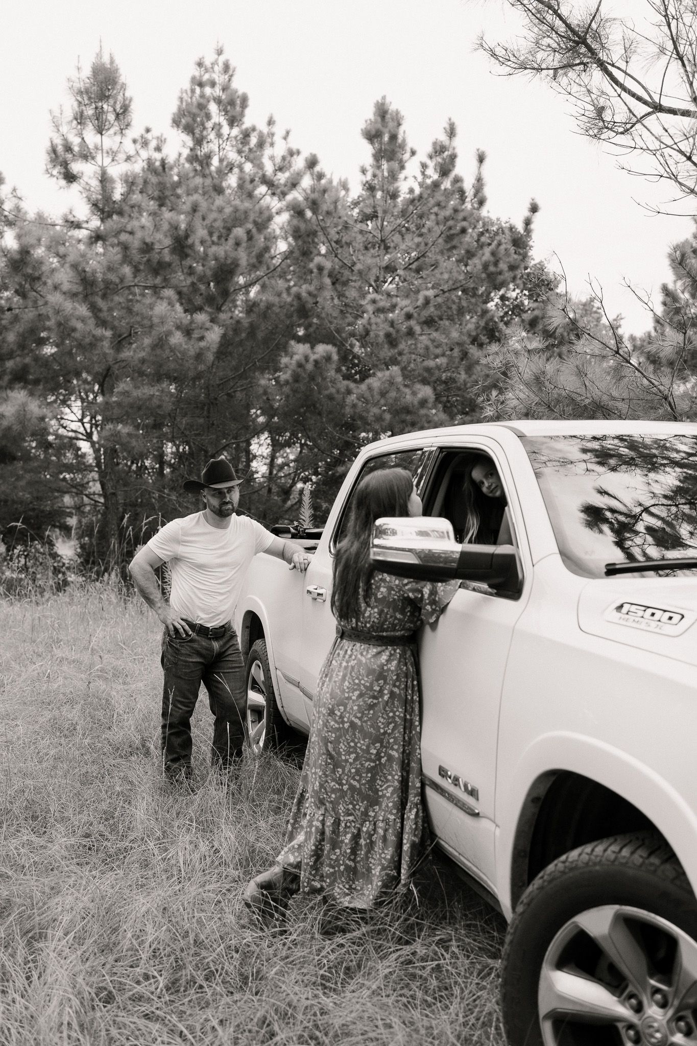 Black and white photo of a man in a cowboy hat leaning against a white pickup truck in a field.