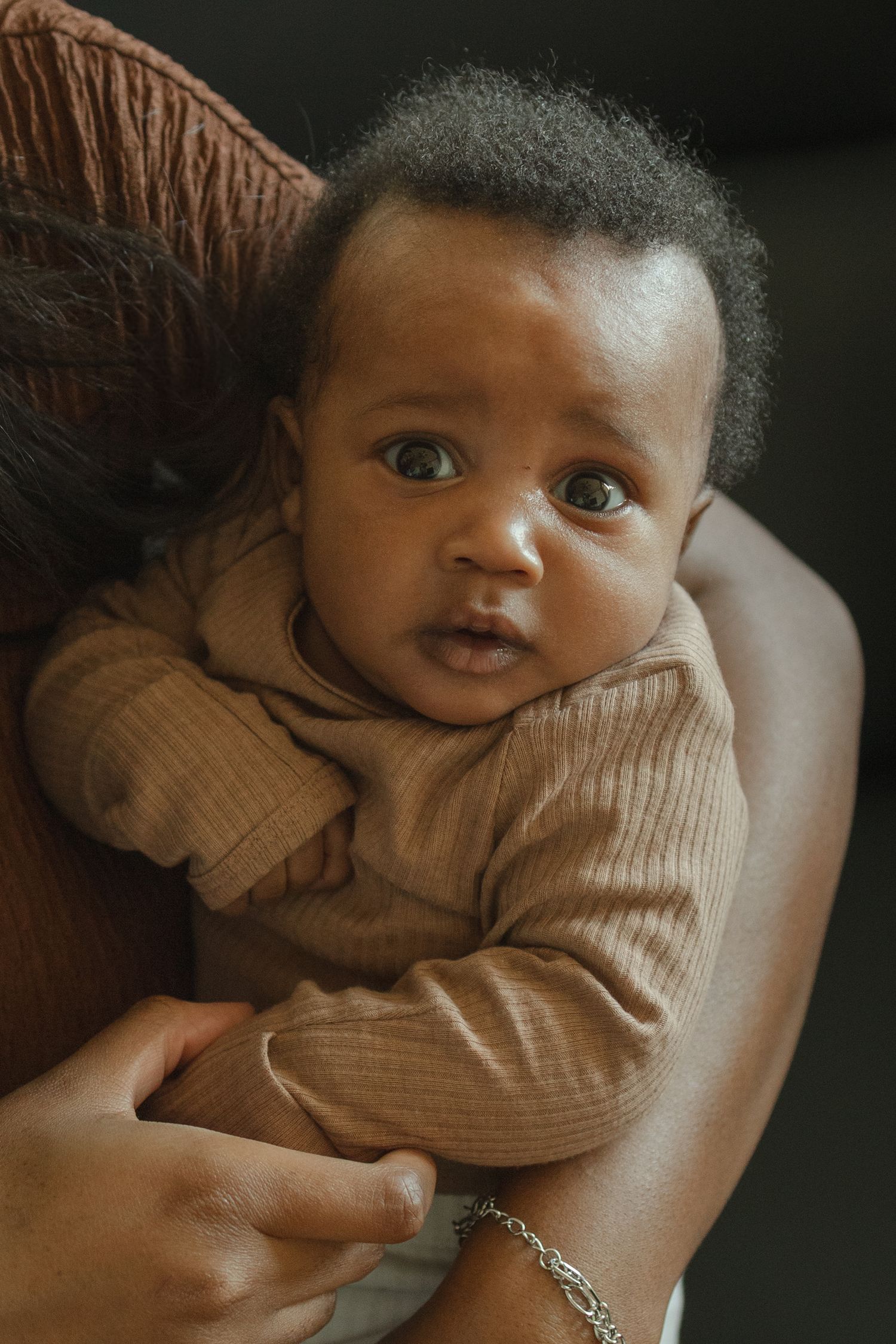 Close-up of alert Black baby in brown onesie being held over parent's shoulder.