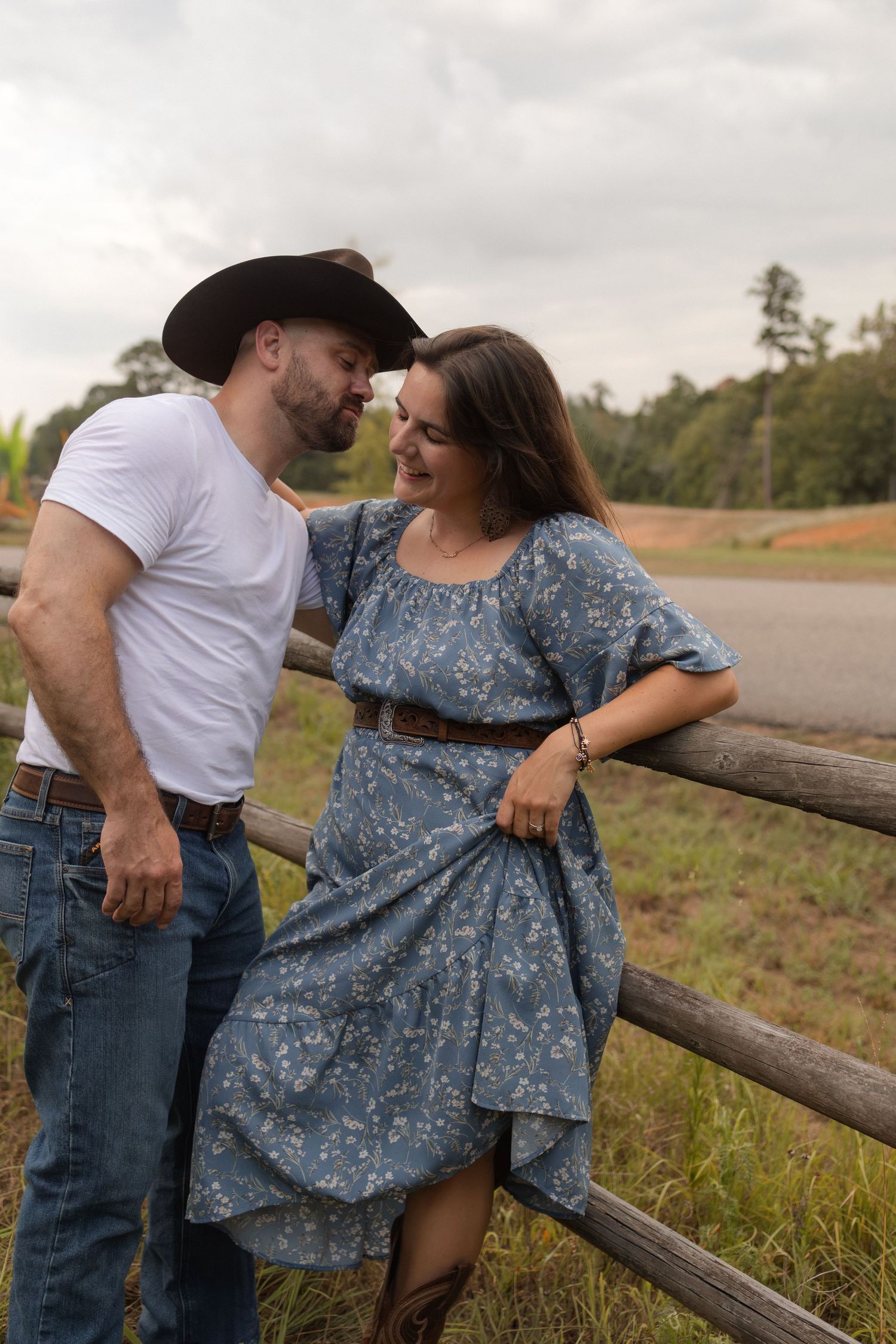 Couple in country style attire leaning against a rustic wooden fence smiling at each other.