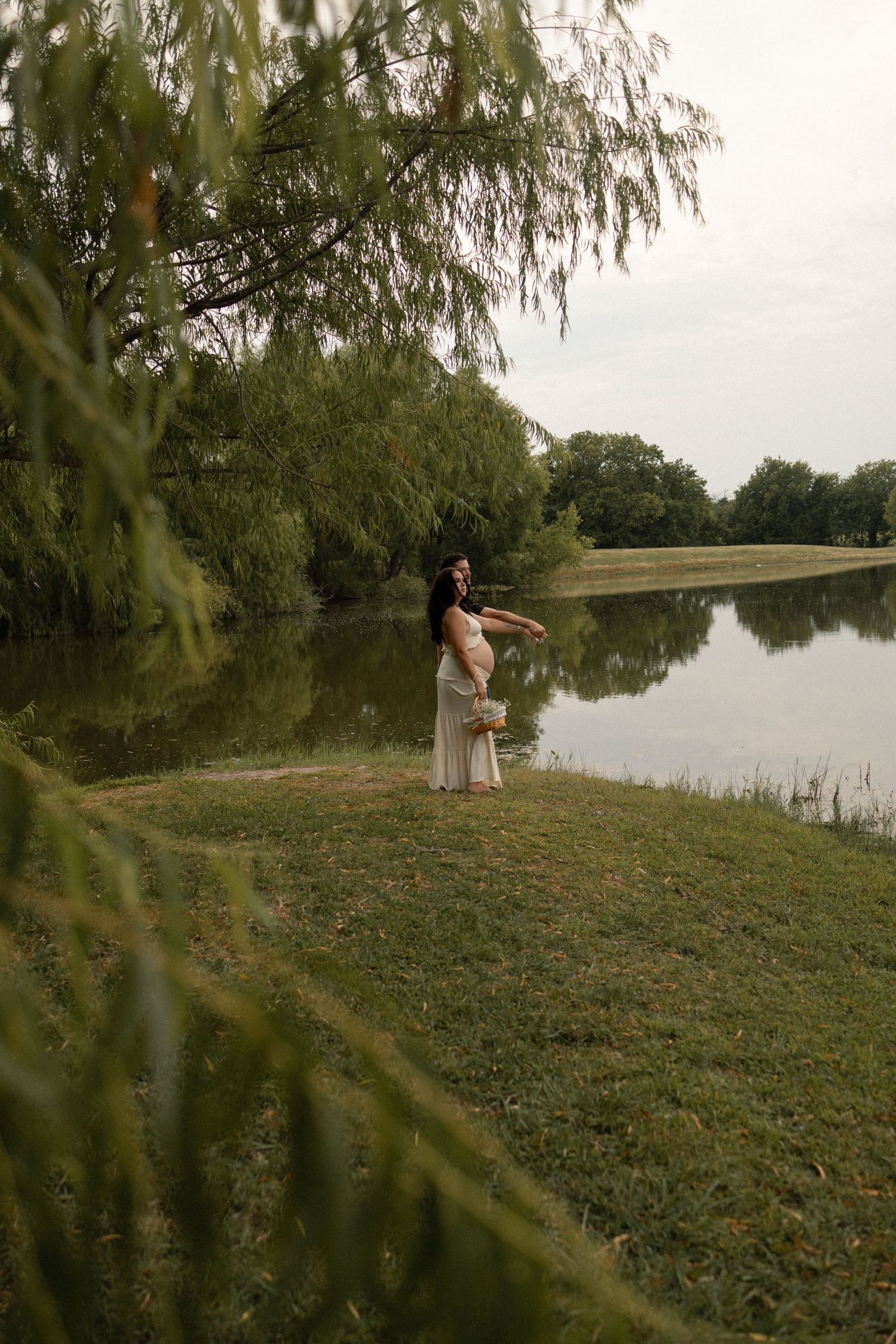 Pregnant woman in a white dress posing gracefully by a reflective lake surrounded by willow trees.
