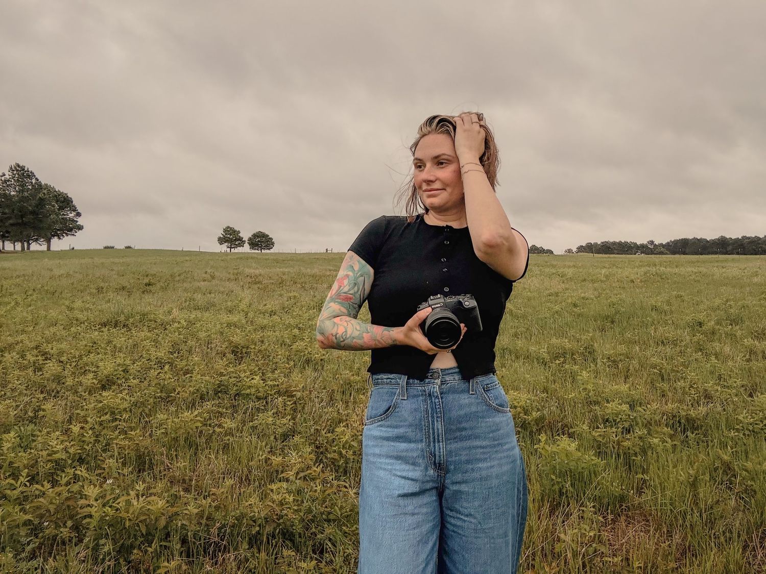Young tattooed woman holding camera in open grassy field, wearing black top and wide-leg jeans, under overcast sky.