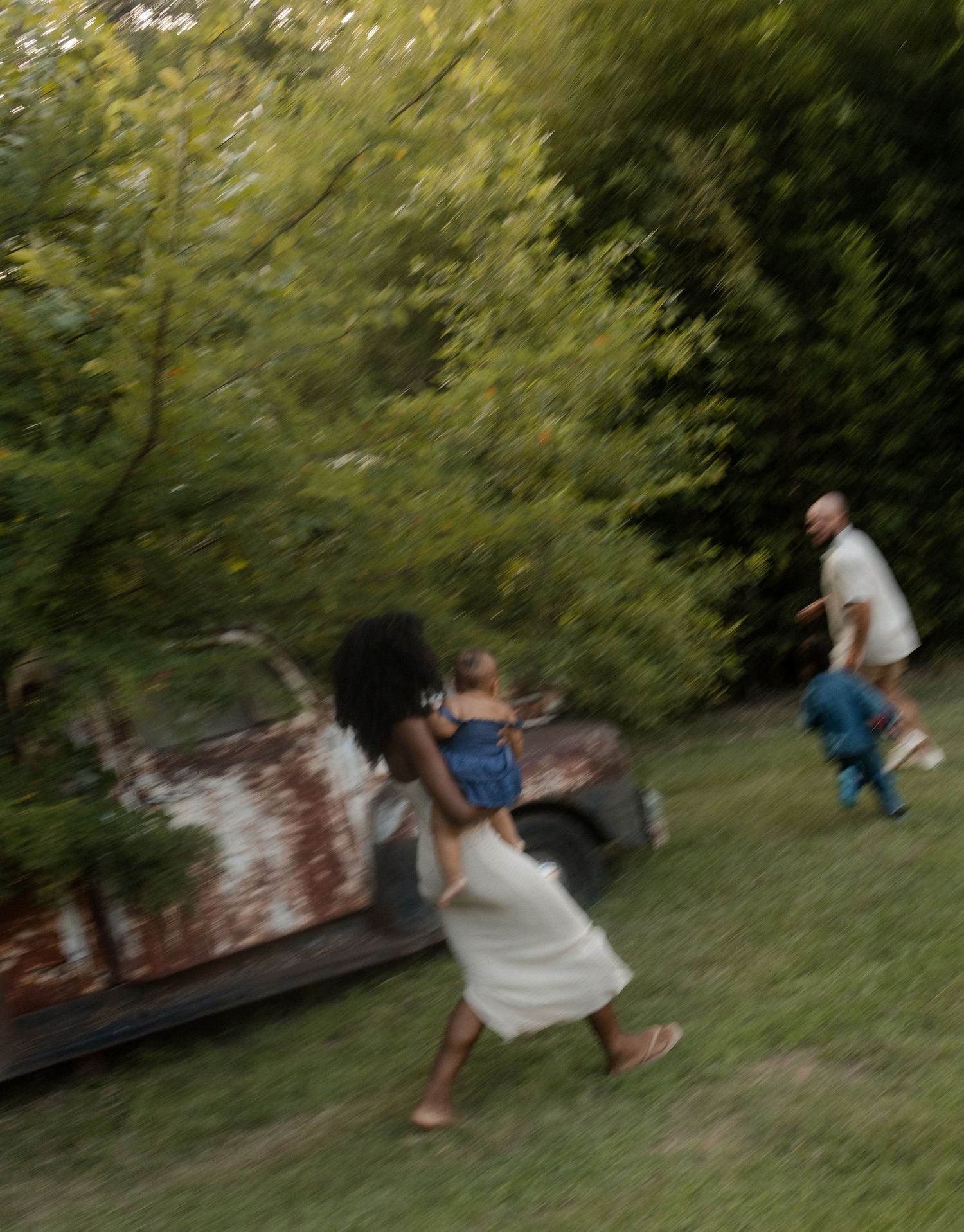 A blurred motion shot of a family playing outdoors near an old rusty vehicle surrounded by green trees.