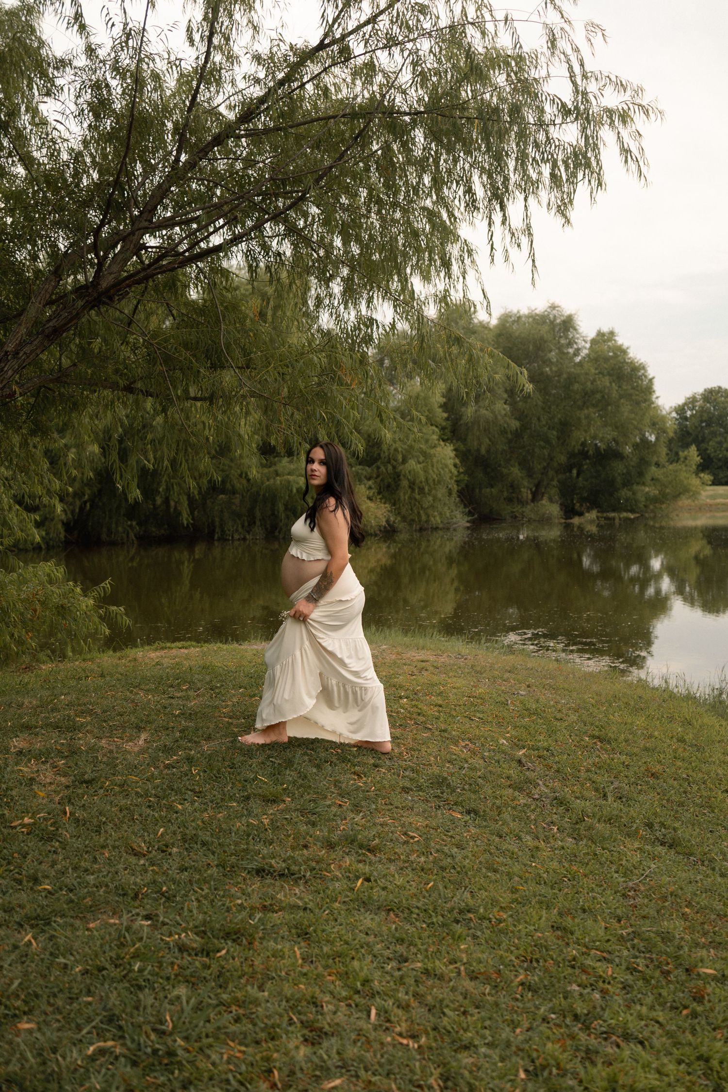 Pregnant woman in white dress standing by tranquil lake under willow tree.