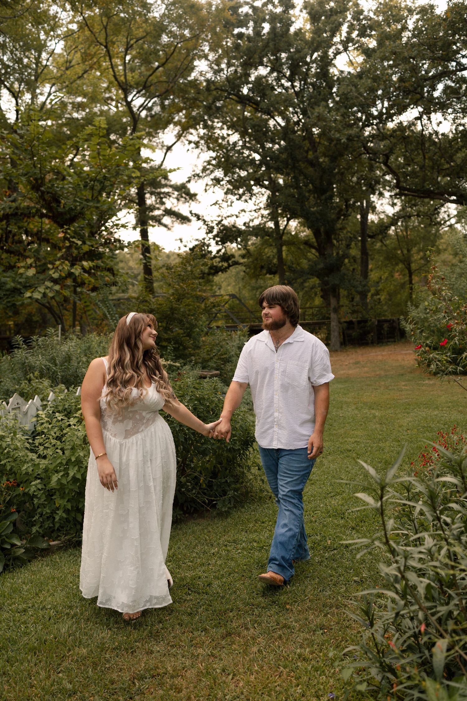 A couple holds hands while walking through a lush garden, woman in white dress, man in white shirt and jeans.