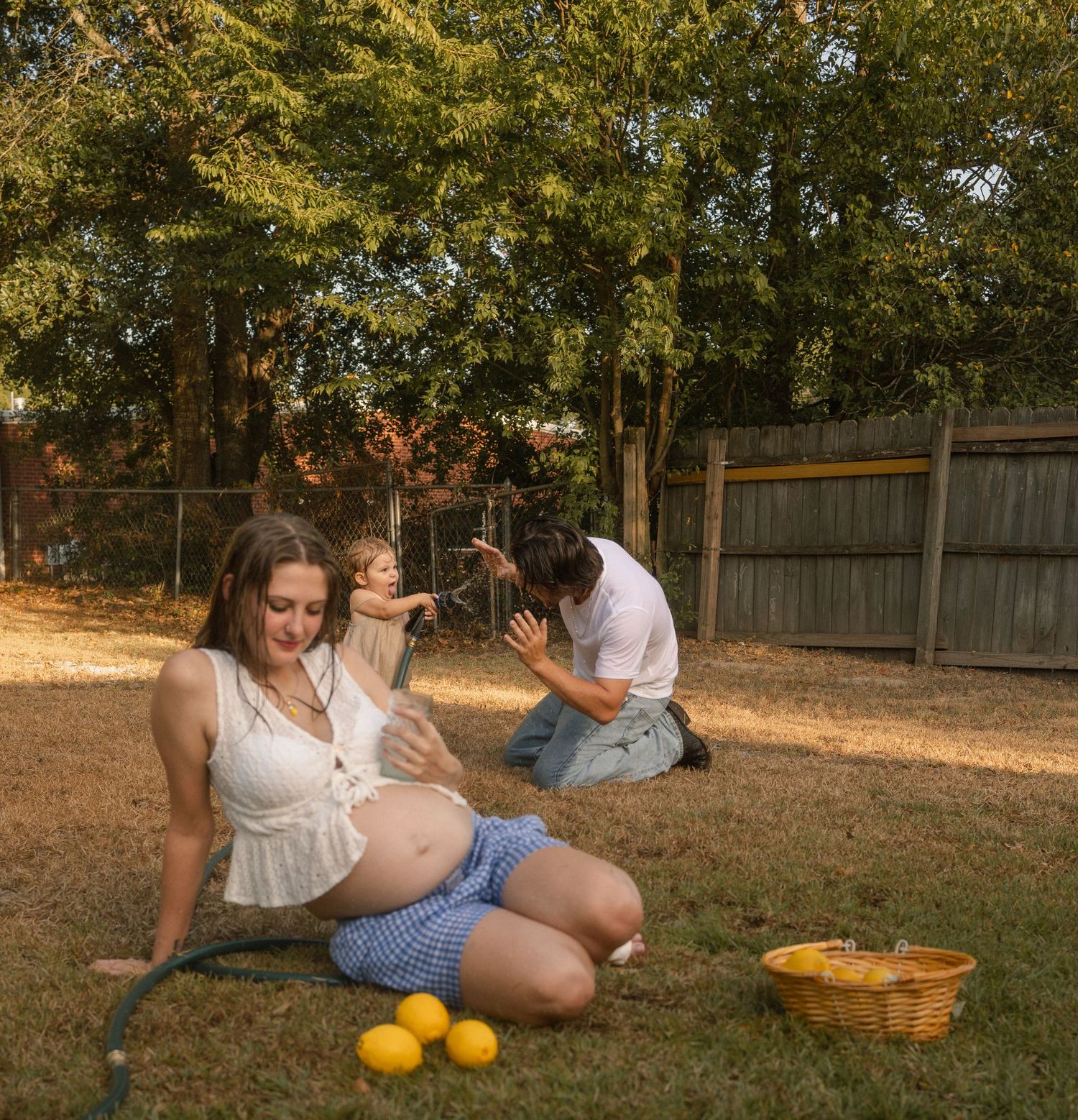 Pregnant woman sitting on grass with partner and toddler playing with a garden hose outdoors.