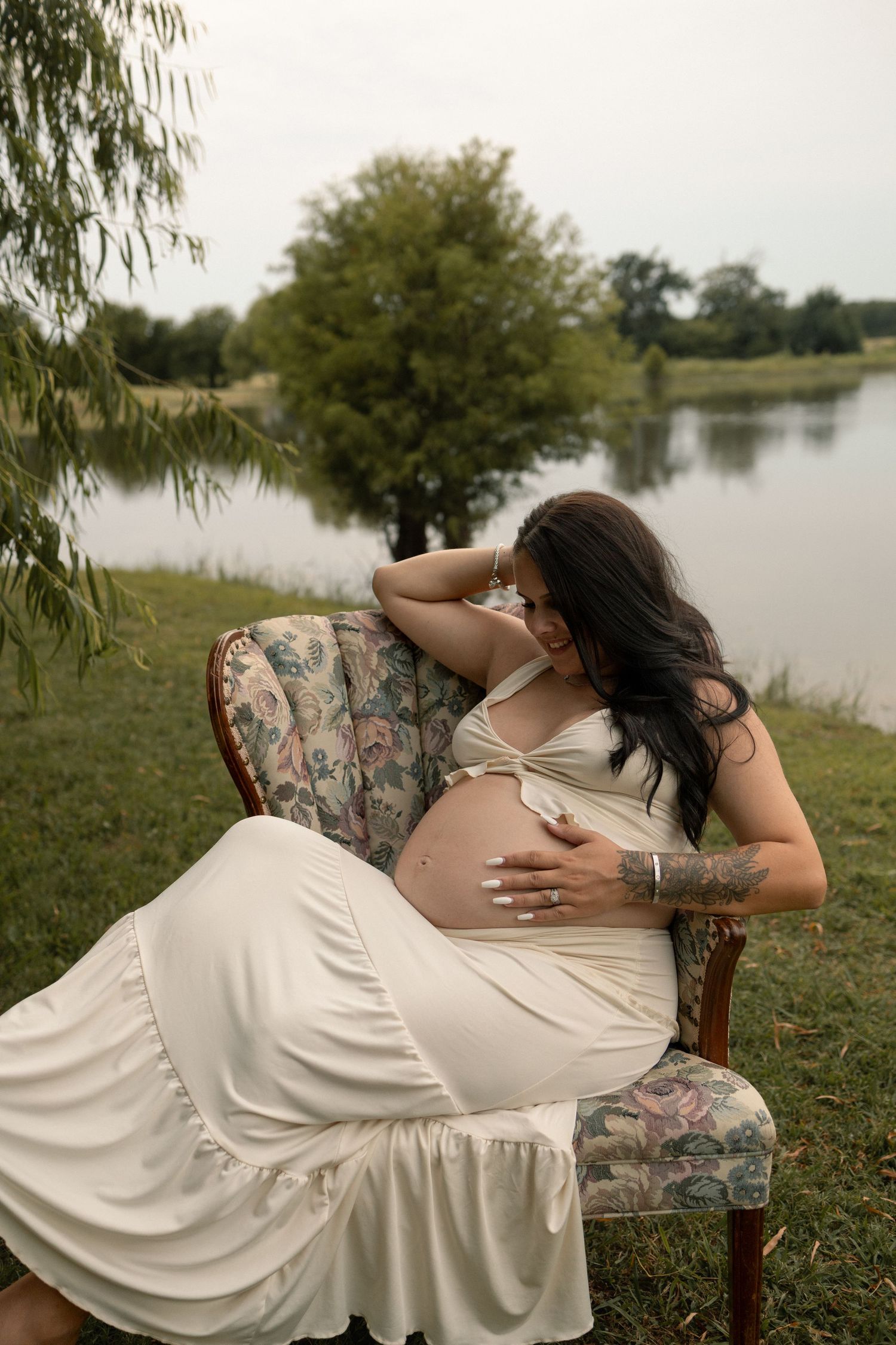 A pregnant woman in a white dress reclines gracefully on a floral chair beside a serene lake.