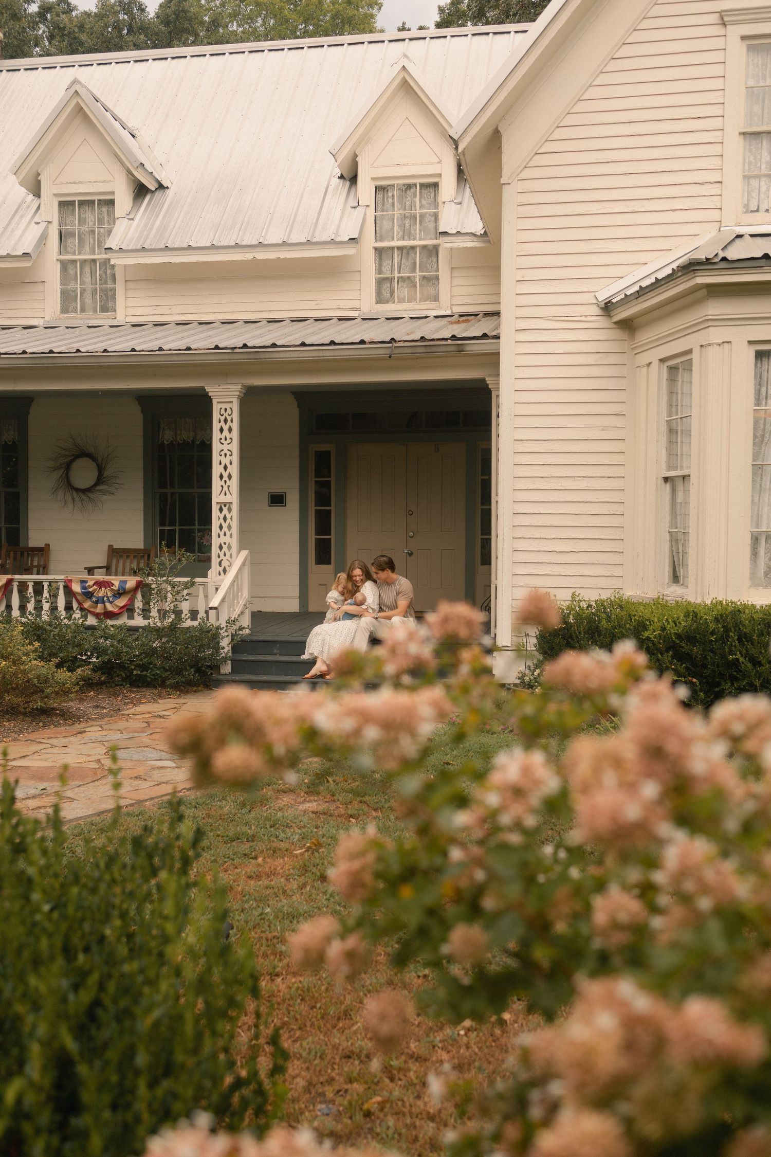 A family sits together on the porch steps of a charming white farmhouse surrounded by blooming gardens.