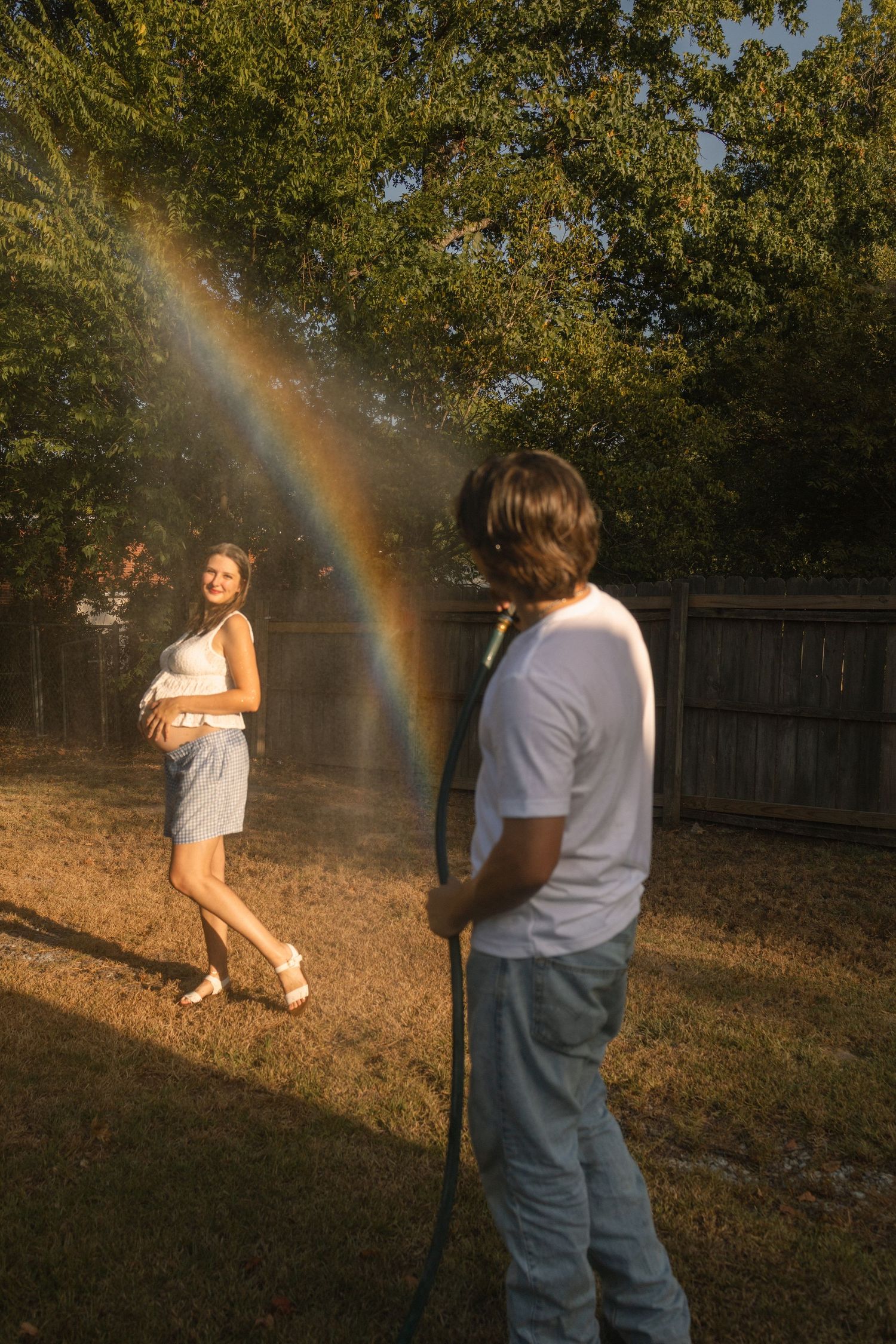 Man spraying garden hose creating a rainbow while pregnant woman smiles in sunny backyard.