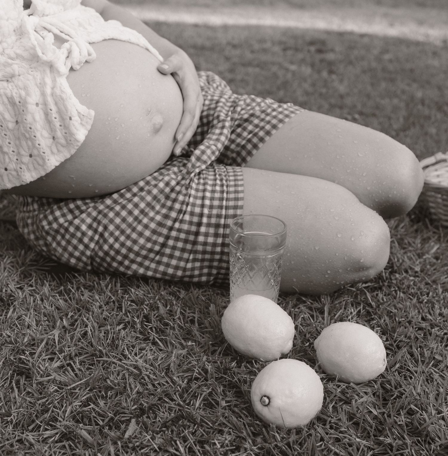 Black and white photo of pregnant woman sitting on grass with lemons and a glass of lemonade nearby.