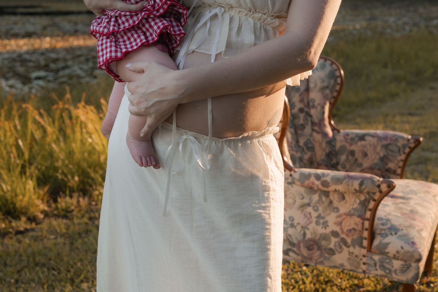 Close-up of a woman in a white dress holding a baby in a red gingham outfit at golden hour.