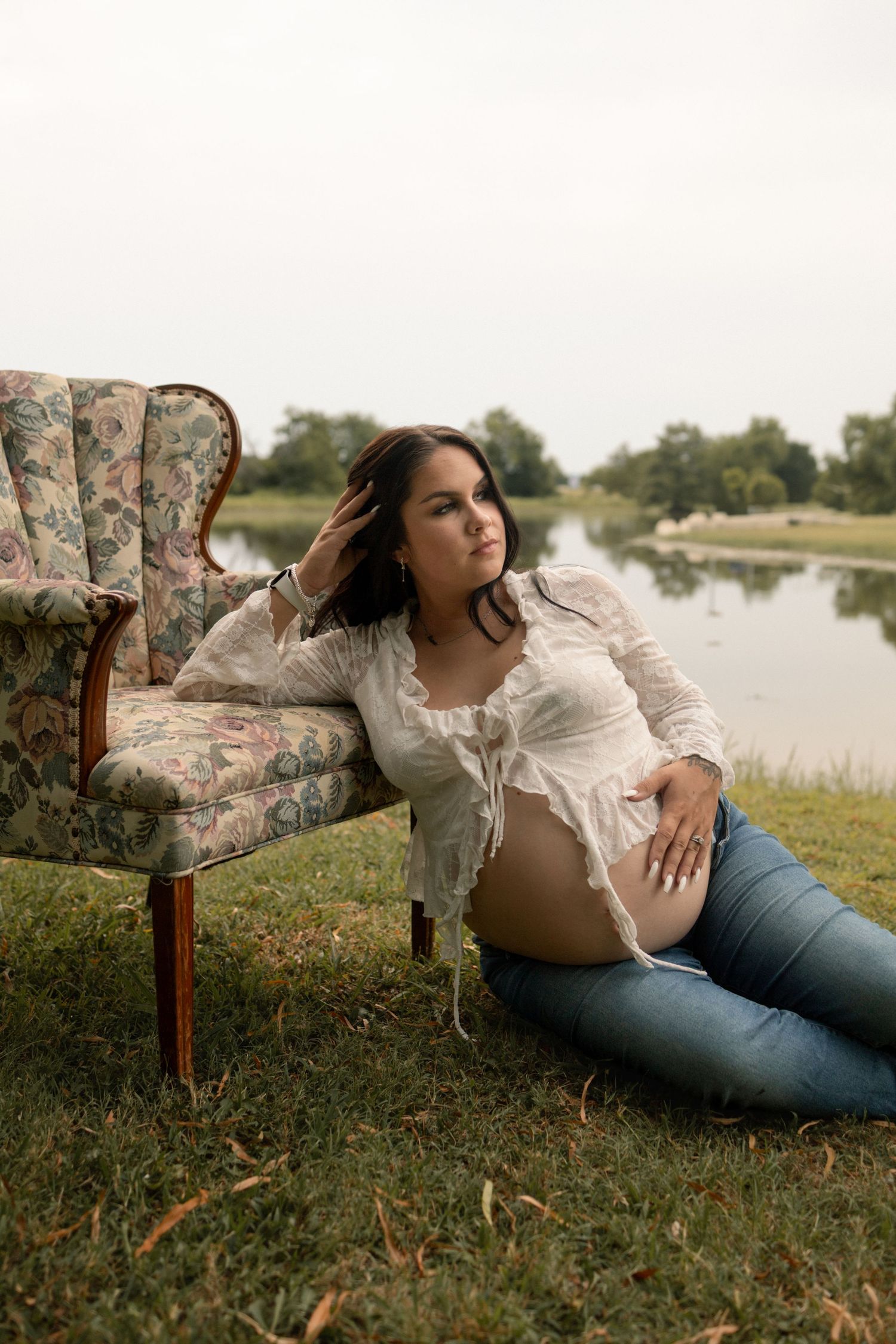 Pregnant woman in white lace top and jeans resting beside vintage floral chair by lake.