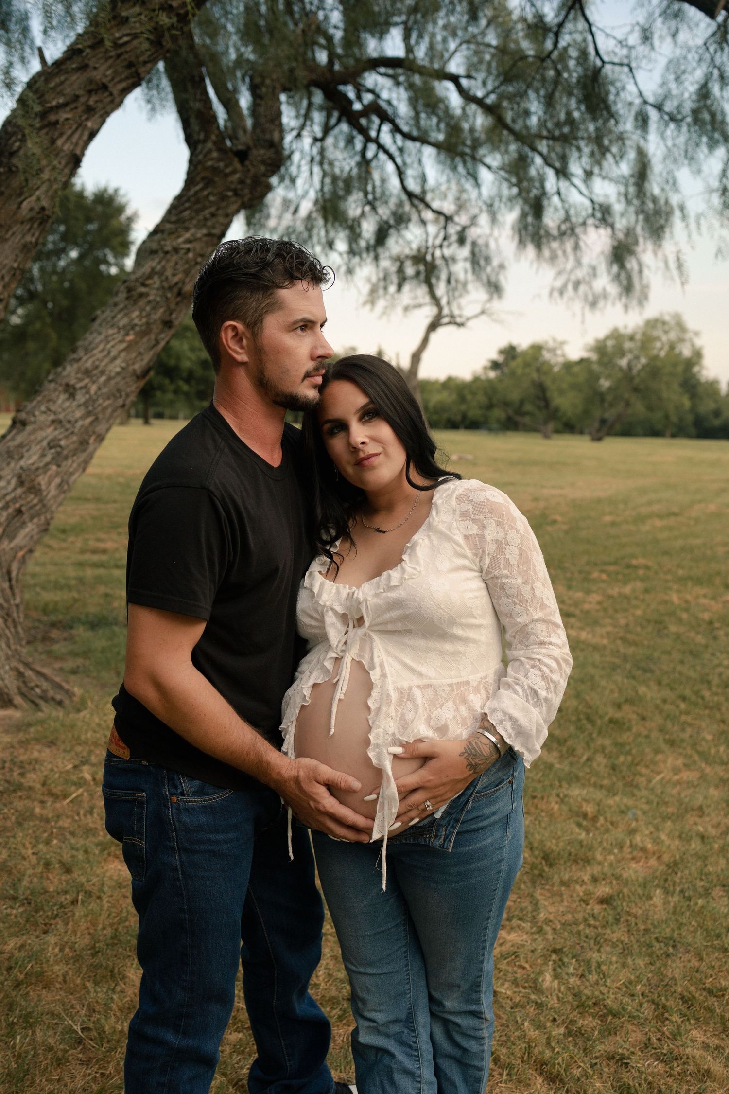 A couple stands under a tree at sunset, man embracing pregnant woman showing her baby bump in a park.