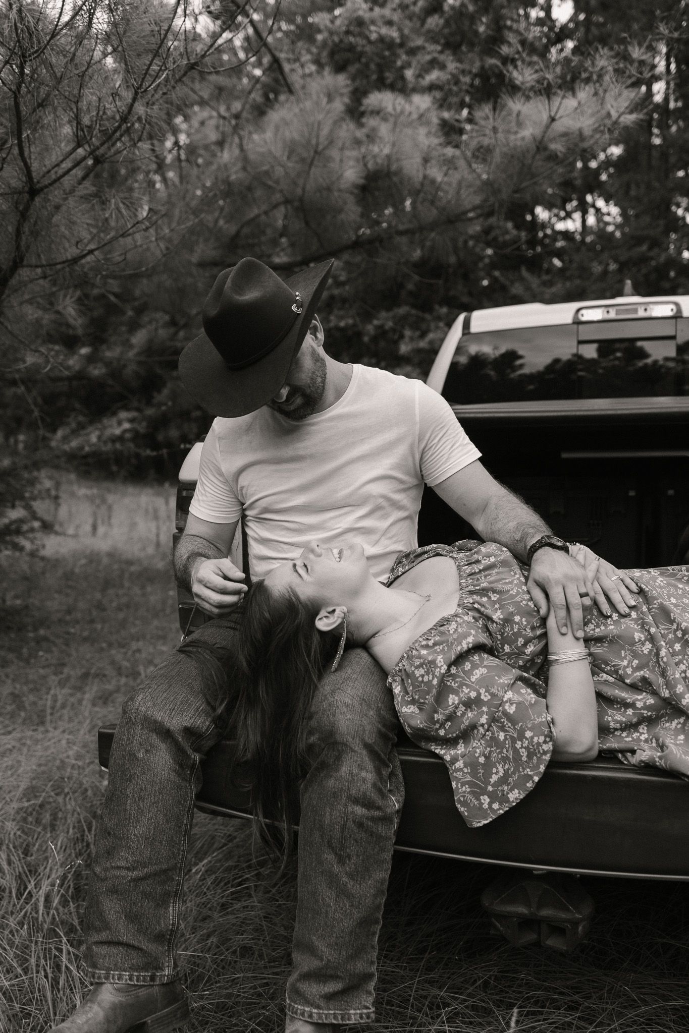 Black and white romantic photo of a couple leaning against a truck tailgate, man in cowboy hat holding woman in floral dress.